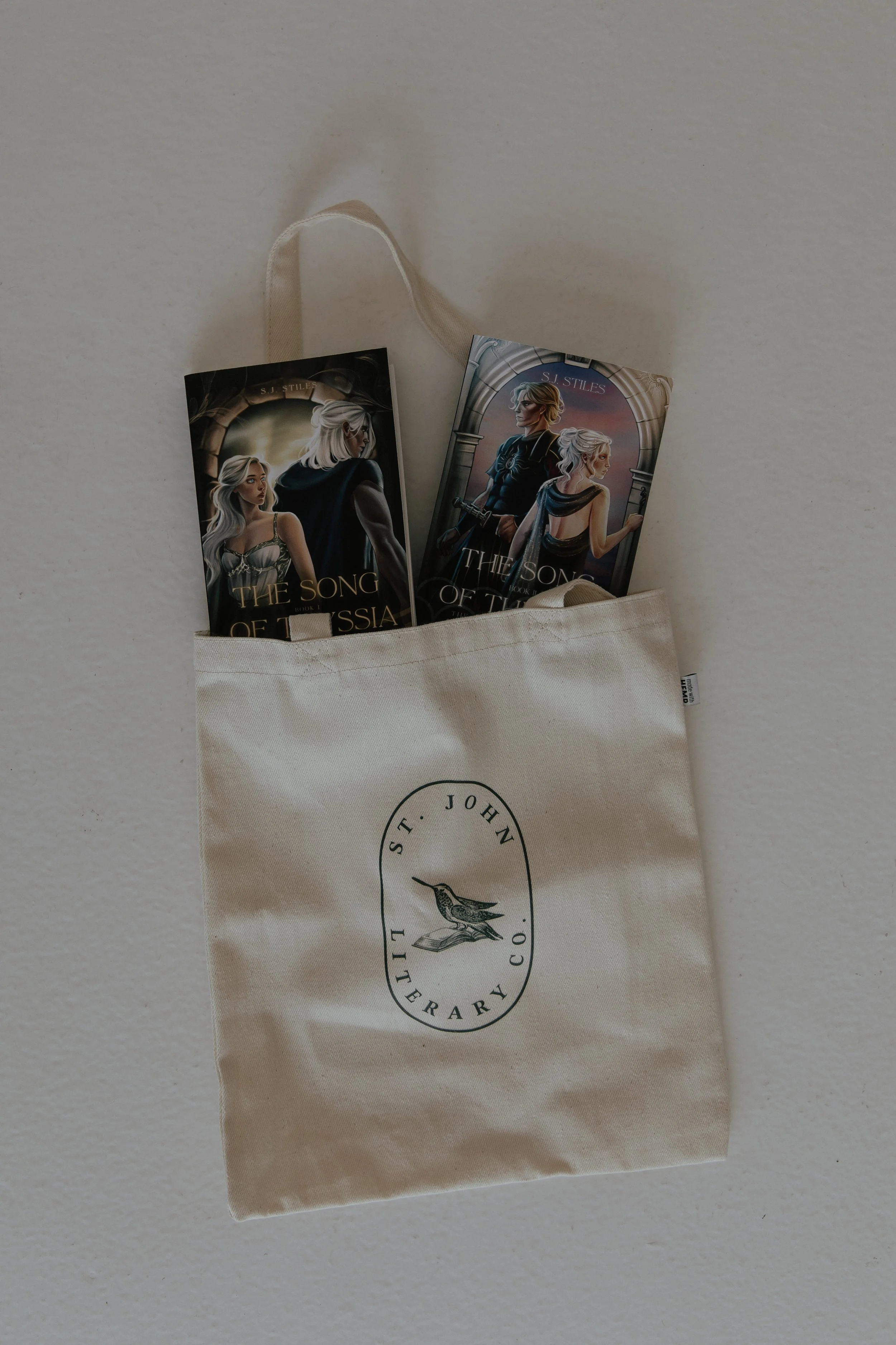 Two book covers from The Song of Ice and Fire series by S. J. Stiles, placed inside a beige tote bag with a logo. The books feature illustrations of characters from the series, with one showing a woman with silver hair and the other a man with blonde hair, set against castle archways.