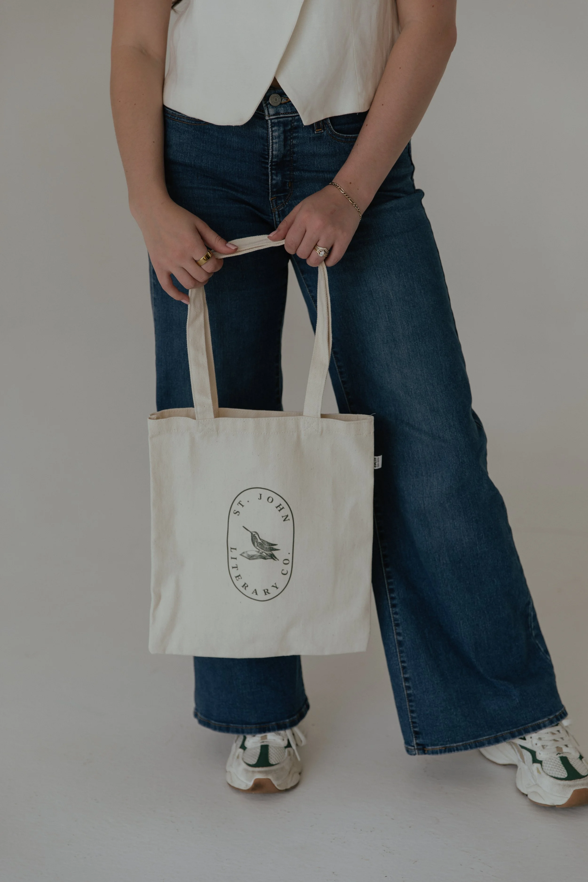 Person wearing a white sleeveless top, blue jeans, and white sneakers with green accents, holding a beige tote bag with a logo that reads 'ST. JOHN'S LIBRARY CO.'