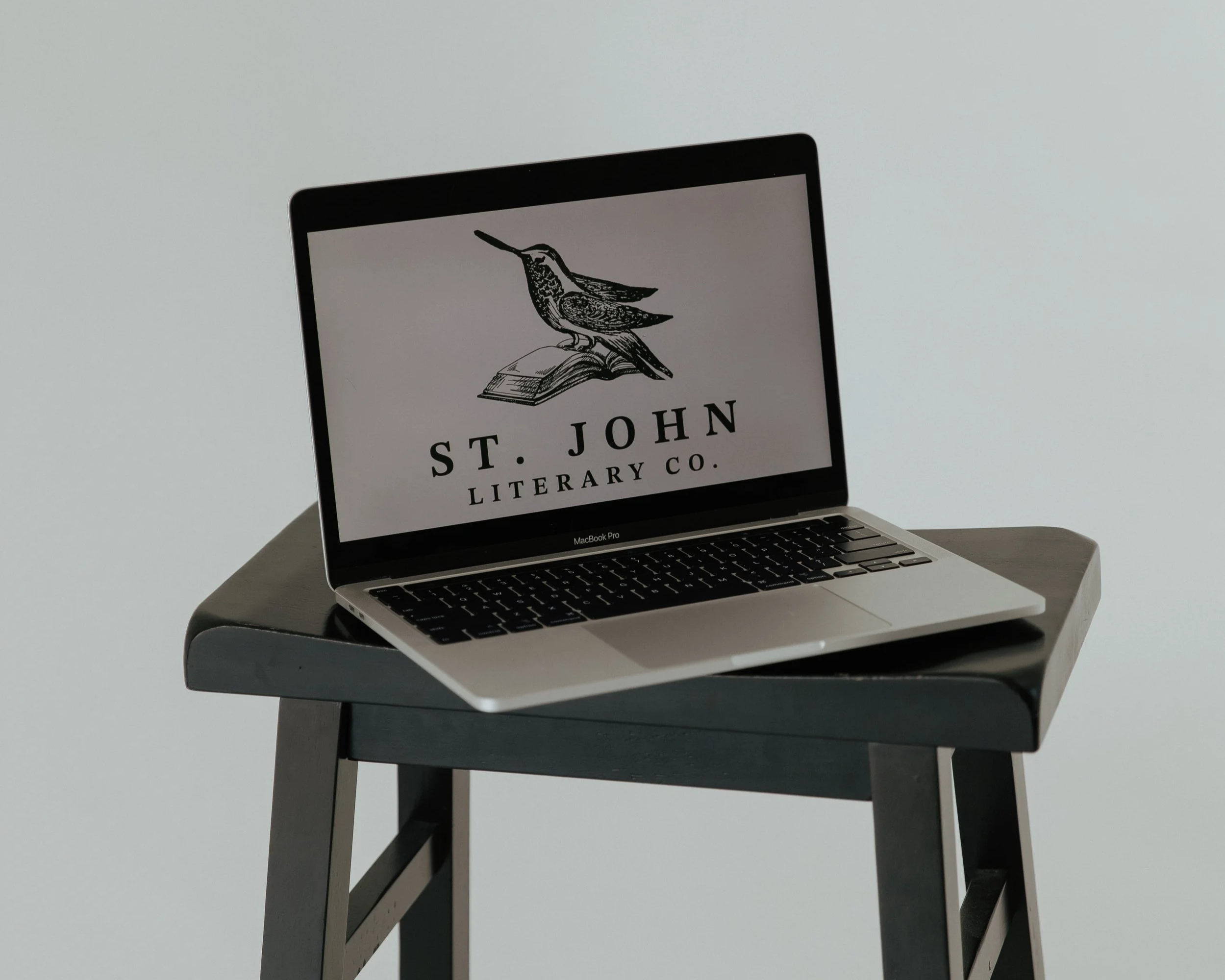 Laptop on gray table displaying a logo with a bird perched on an open book and the text 'St. John Literary Co.'