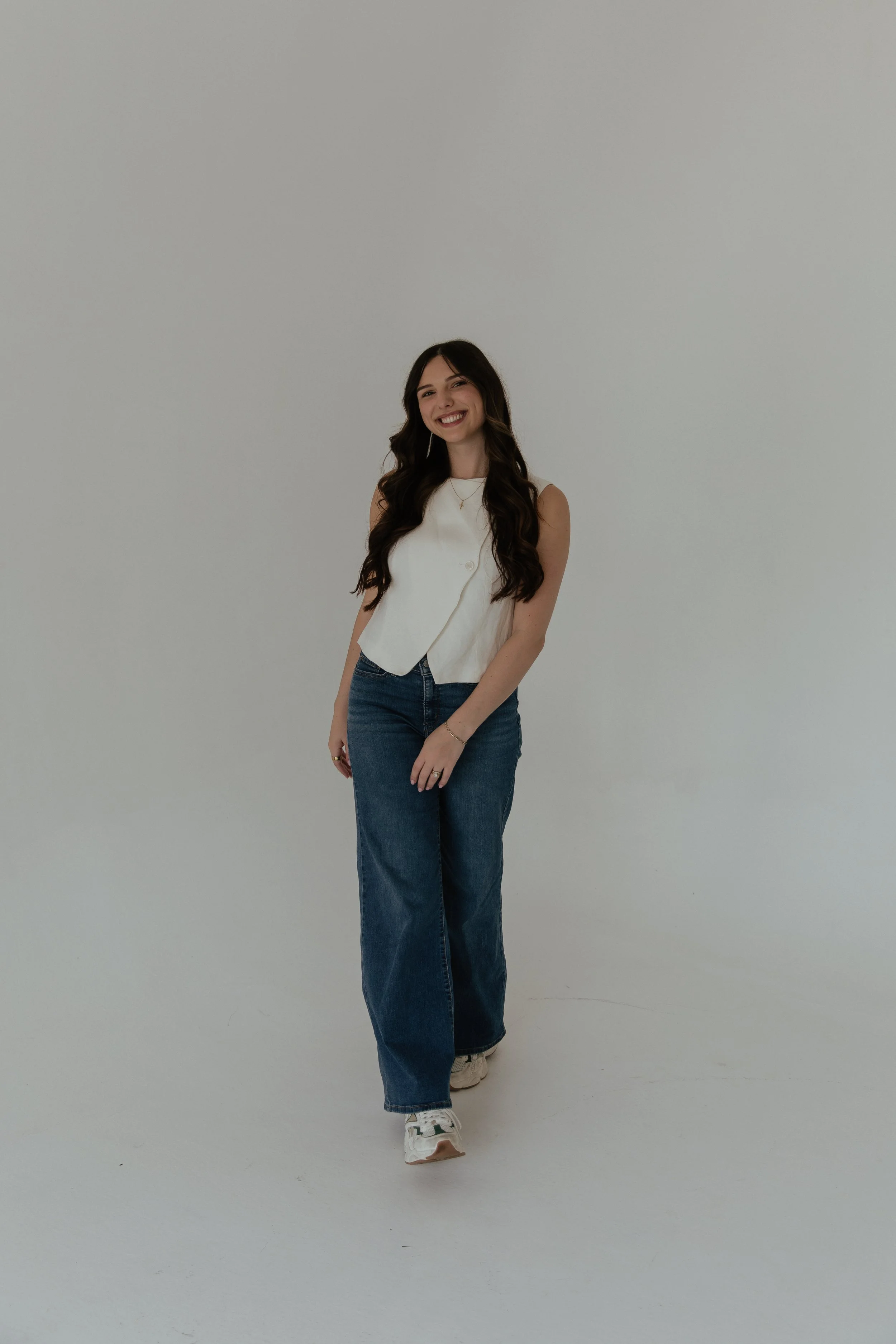 A young woman with long dark hair smiling and wearing a sleeveless white top and blue jeans, standing against a plain white background.