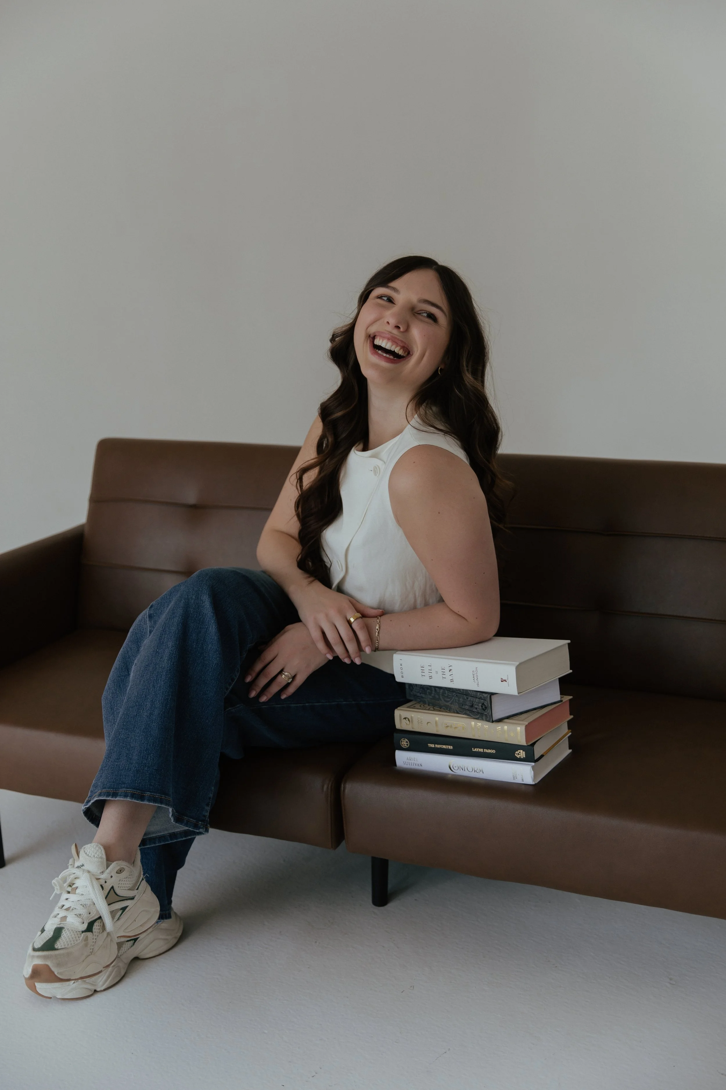 A smiling woman with long dark wavy hair sitting on a brown couch, wearing a white sleeveless top, blue jeans, and white sneakers, with a stack of books beside her.