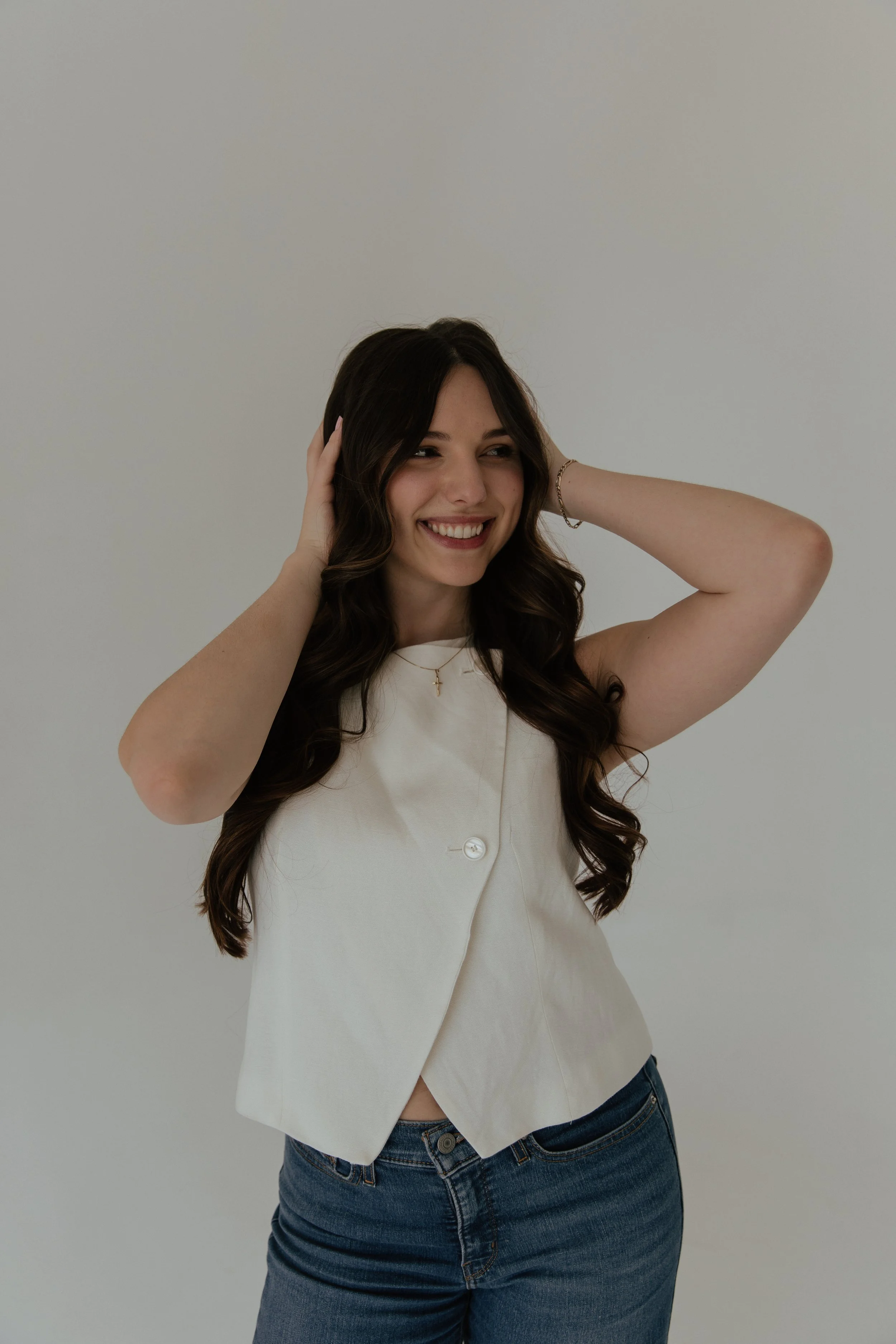 Young woman with long, wavy brown hair, smiling, holding her hands to her head, wearing a white sleeveless top and blue jeans, standing against a plain light-colored background.