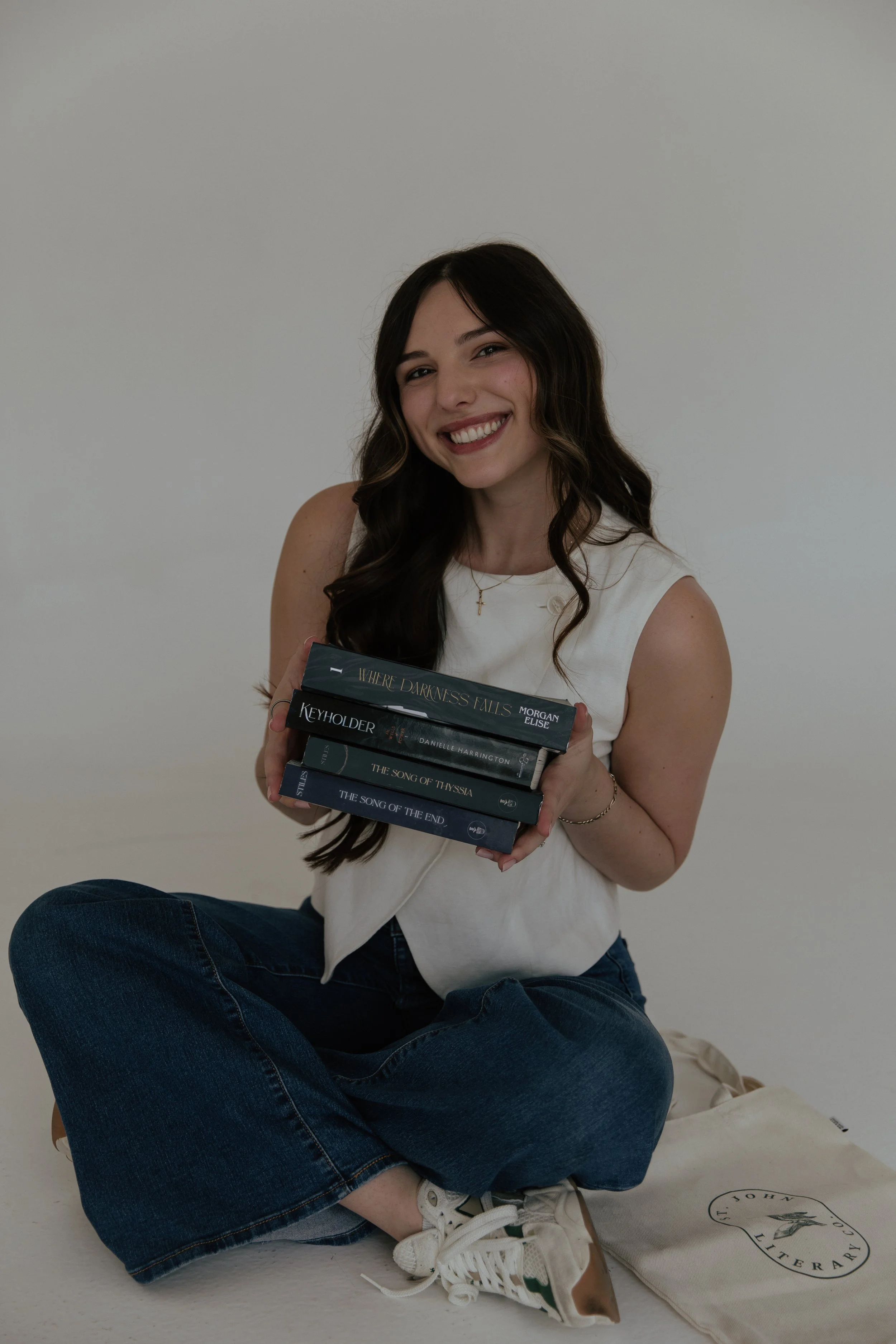 A young woman with long dark hair, wearing a white sleeveless top and jeans, is sitting cross-legged on the floor, smiling and holding several books. She has a gold cross necklace and is sitting in front of a plain white background.