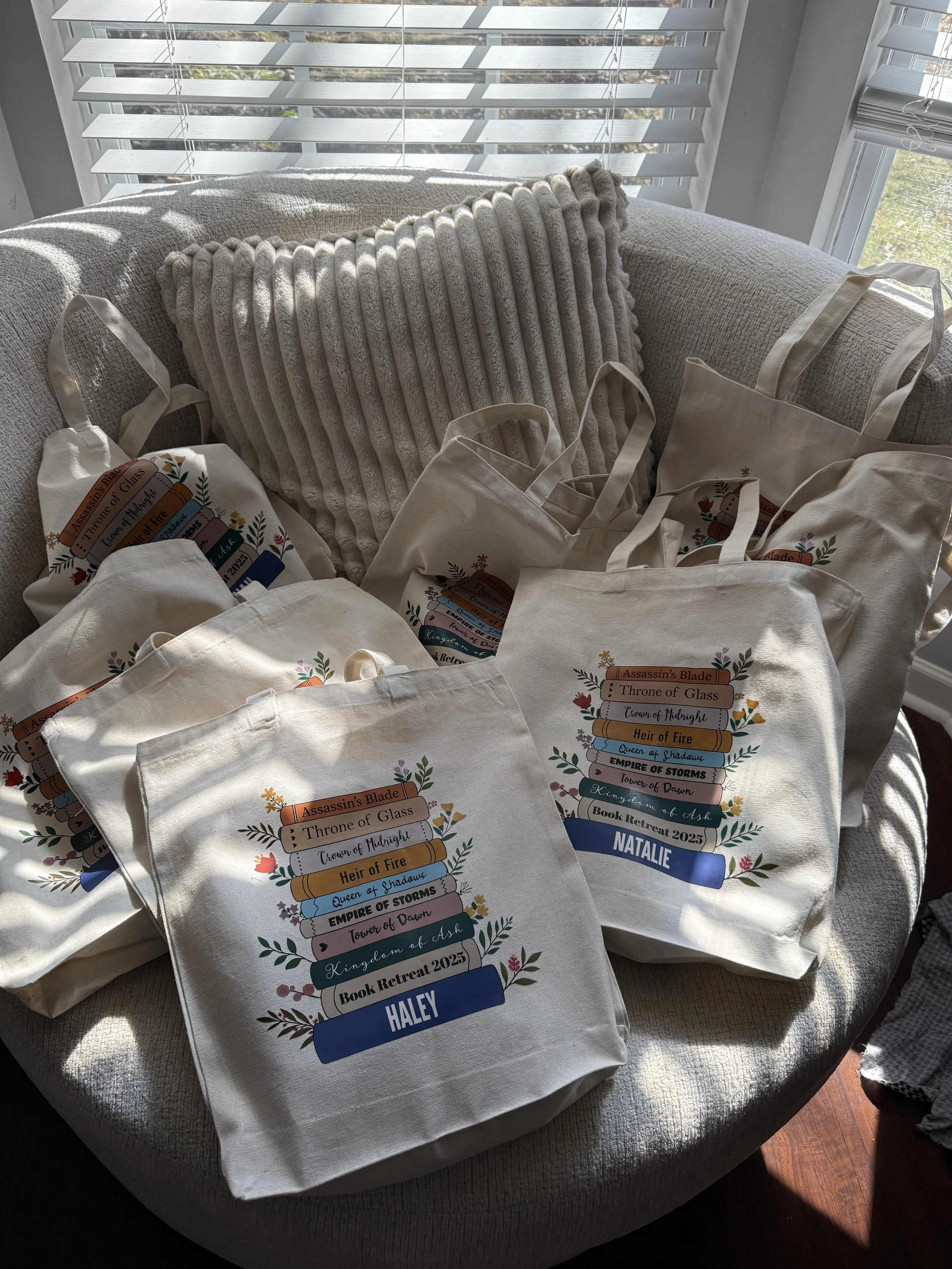 Several tote bags with colorful book-themed designs and individual names, placed on a sofa near window blinds, with sunlight casting shadows.