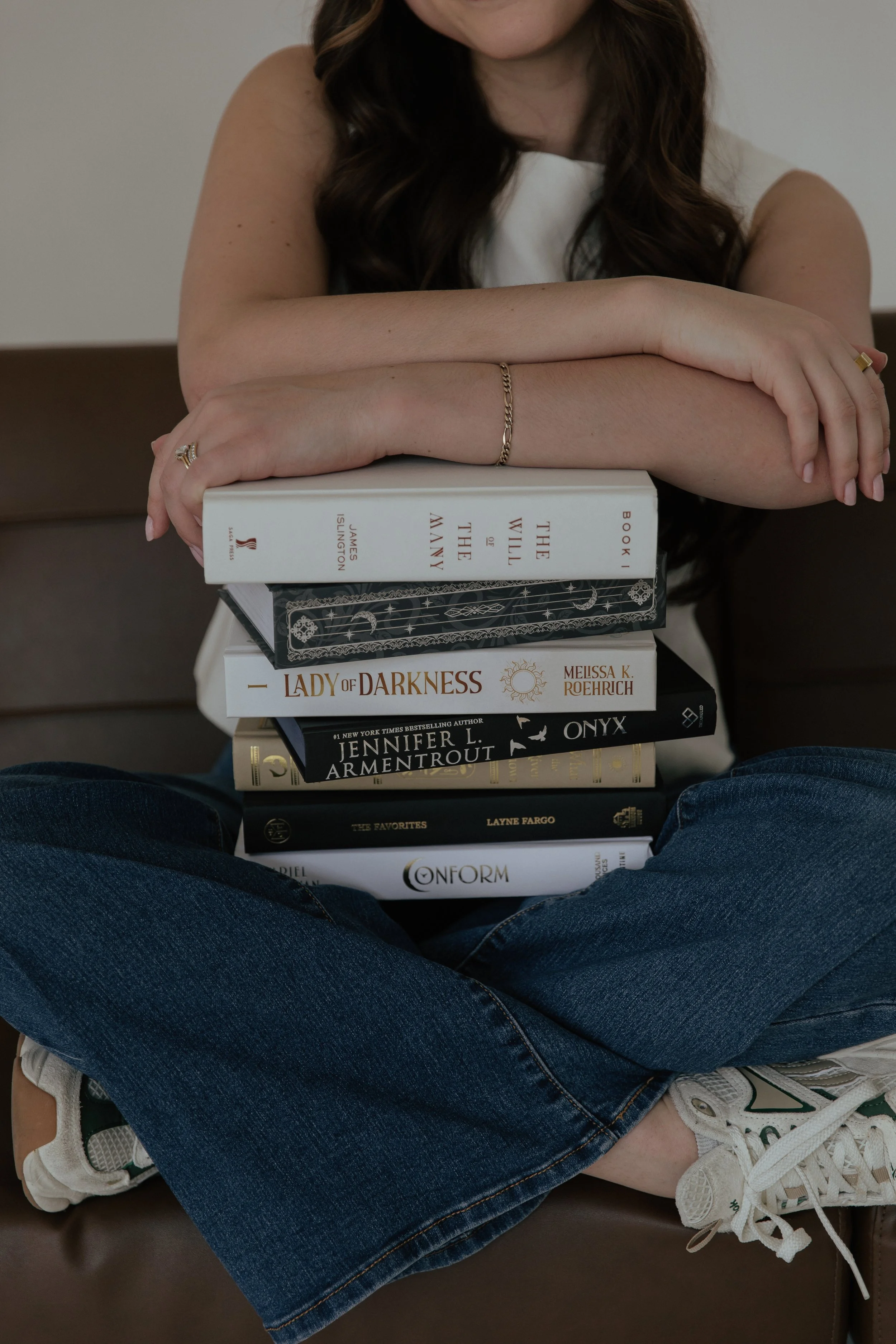 A woman with brown hair sitting cross-legged on a chair, resting her arms on a stack of books, wearing white sneakers, blue jeans, and a white top.