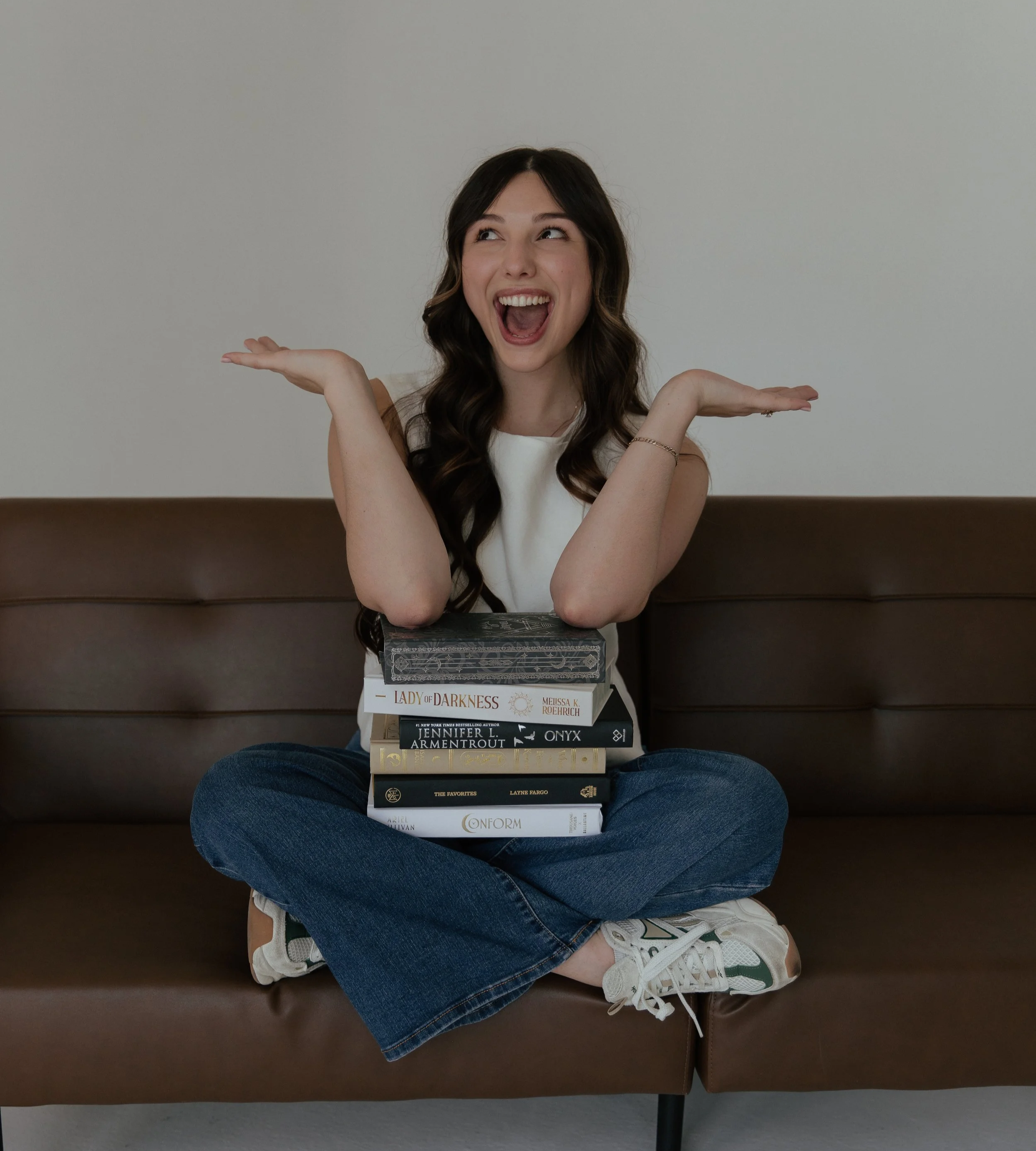A young woman with long dark hair, wearing a sleeveless top, is sitting cross-legged on a brown couch, smiling and gesturing with her hands. There are several books stacked on her lap, including titles like 'Lady of Darkness' and 'Conform,' and she is wearing white sneakers with green accents.