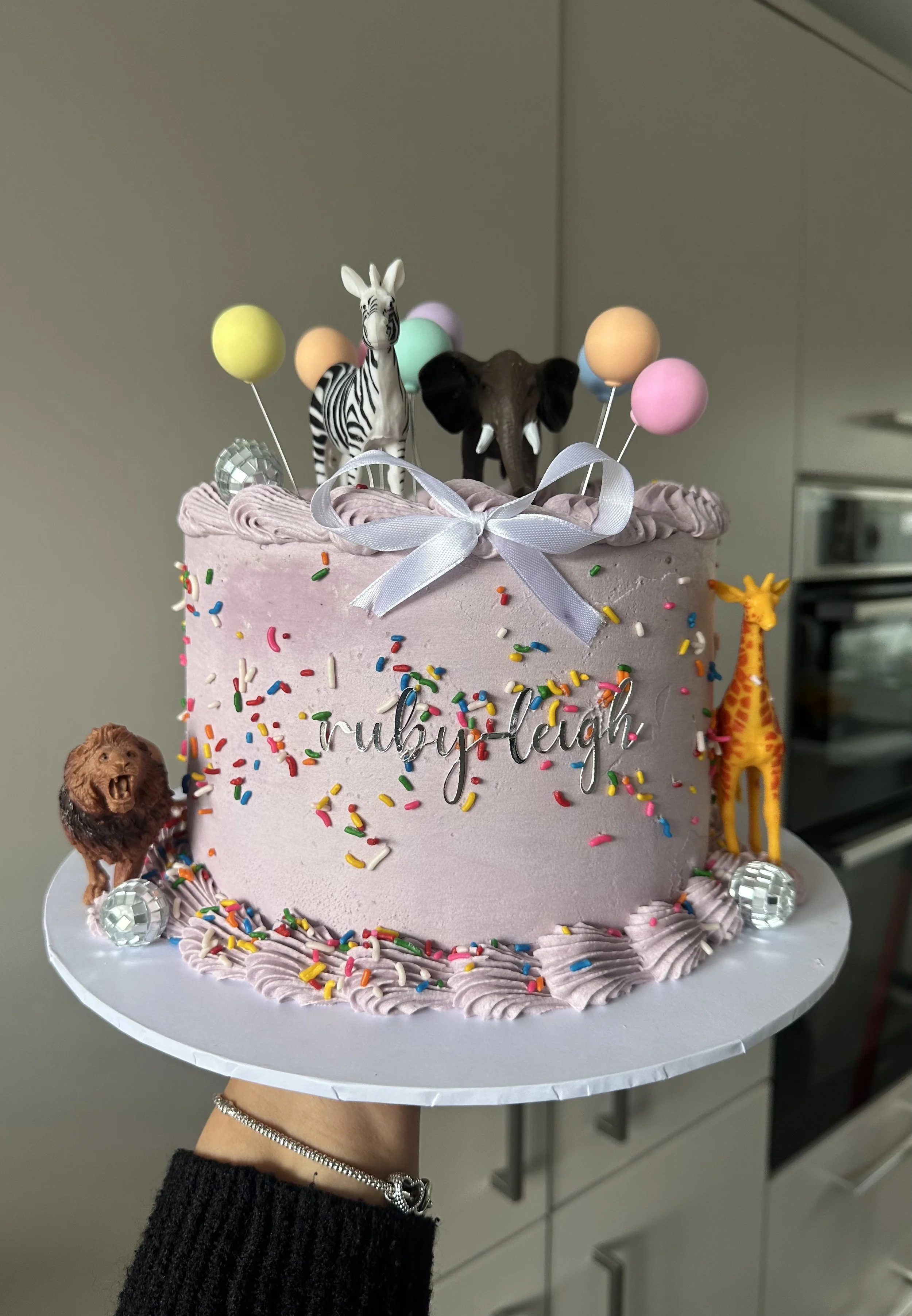 A pink birthday cake with colorful sprinkles, decorated with animal figurines including a zebra, elephant, lion, and giraffe, topped with balloons, a silver ribbon, and a white ribbon tied in a bow, with the name 'ruby-hah' written on the front.