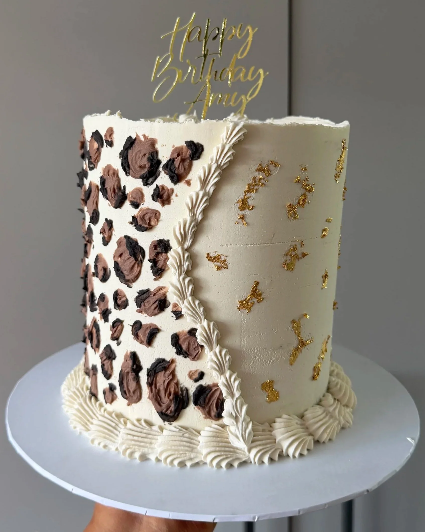 A two-tier birthday cake with white frosting decorated with gold leaf accents, black and brown chocolate frosting toppers resembling abstract flowers, and a gold "Happy Birthday Aray" cake topper. The cake is placed on a white round cake board.