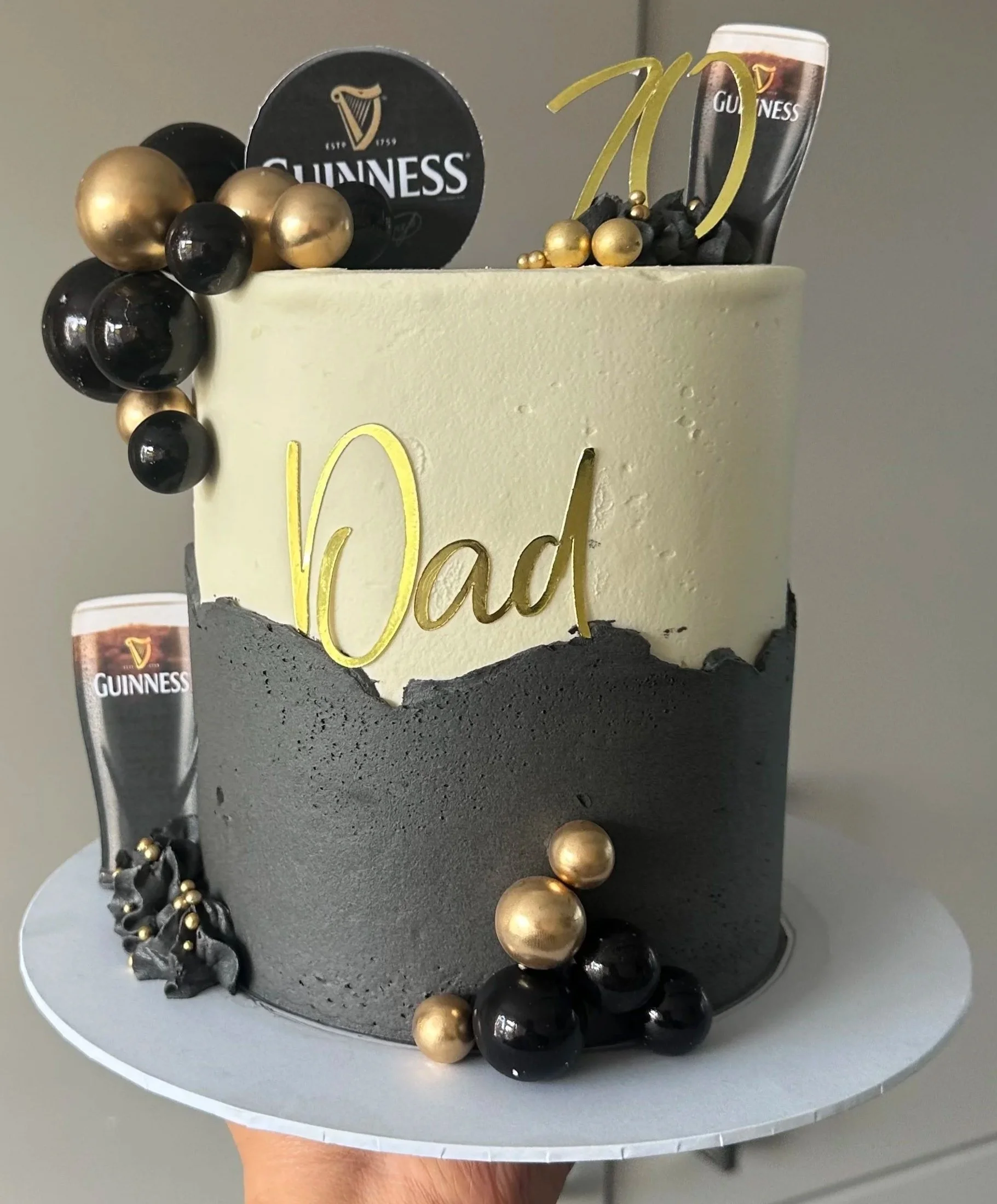 A cake decorated with black, white, and gold elements, featuring a Guinness logo, gold and black spherical decorations, and a gold '70' topper, placed on a white cake stand.