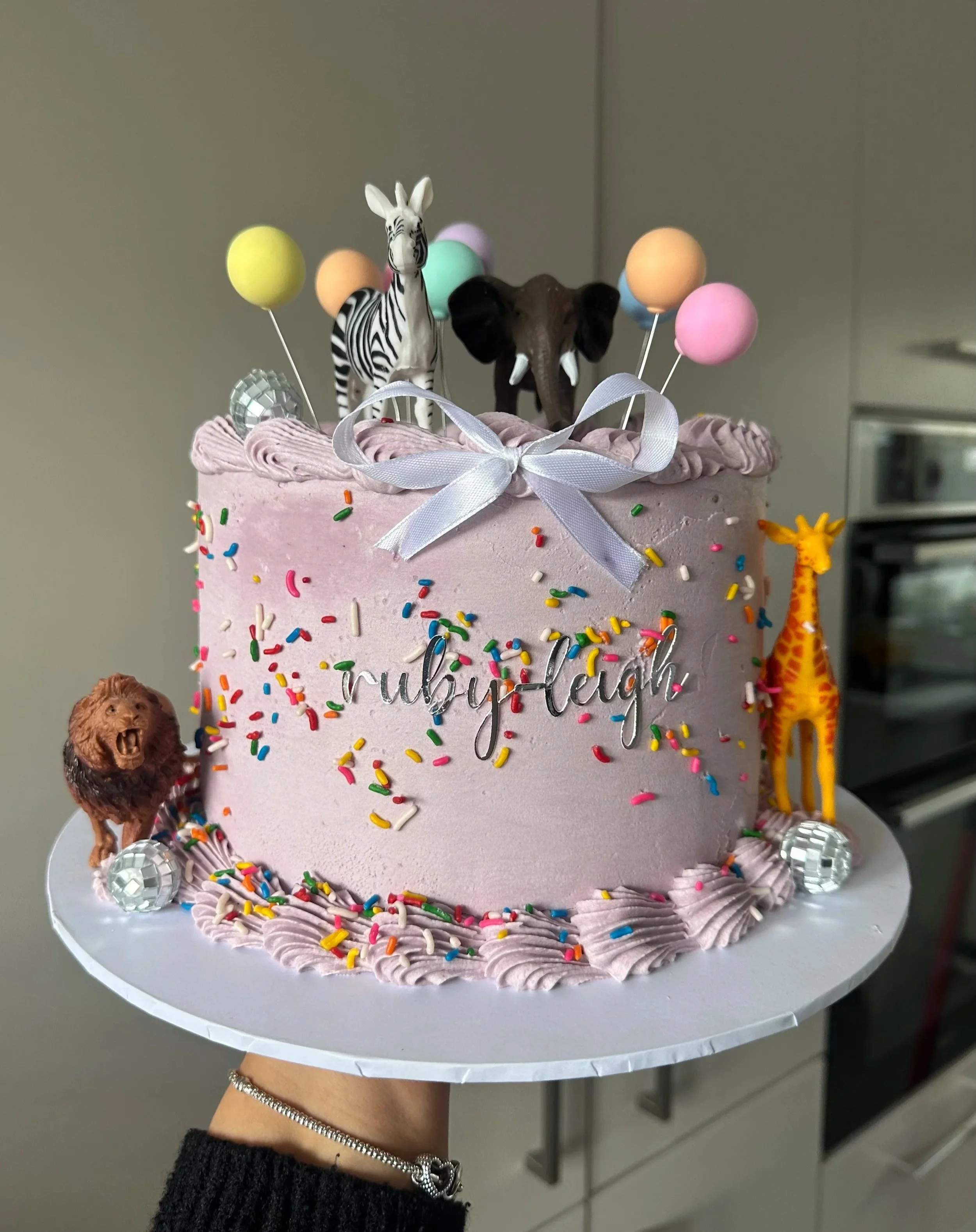 A pink birthday cake decorated with multicolored sprinkles, topped with a white satin bow, toy animal figurines including a zebra and an elephant, and pastel-colored balloons on sticks. The cake is on a white cake stand held by a person with a silver bracelet.