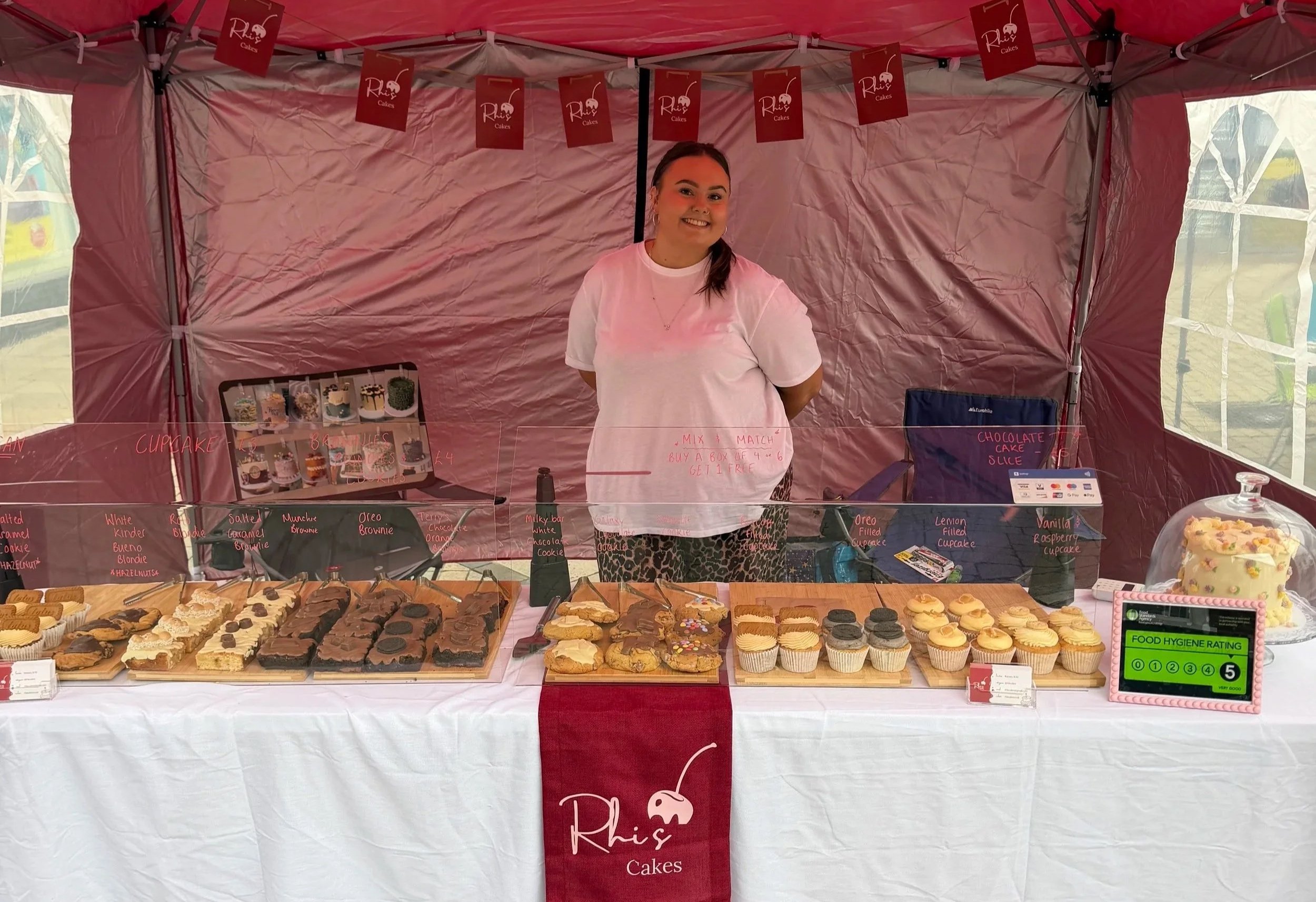 A woman with dark hair and earrings smiling behind a stall filled with various decorated baked goods such as cupcakes and cookies, at an outdoor market with a red canopy and signs reading 'Rhis Cakes'.