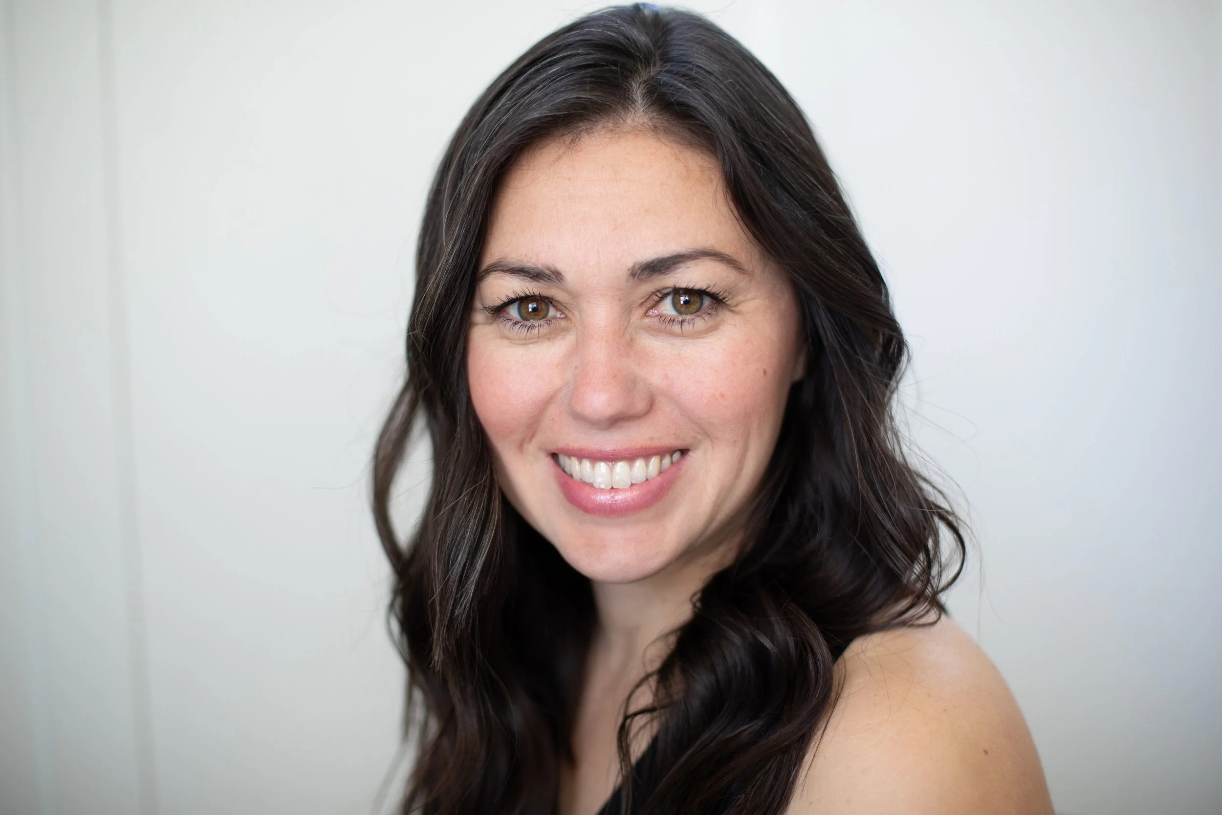 Close-up portrait of a smiling woman with long dark wavy hair and brown eyes against a plain light background.