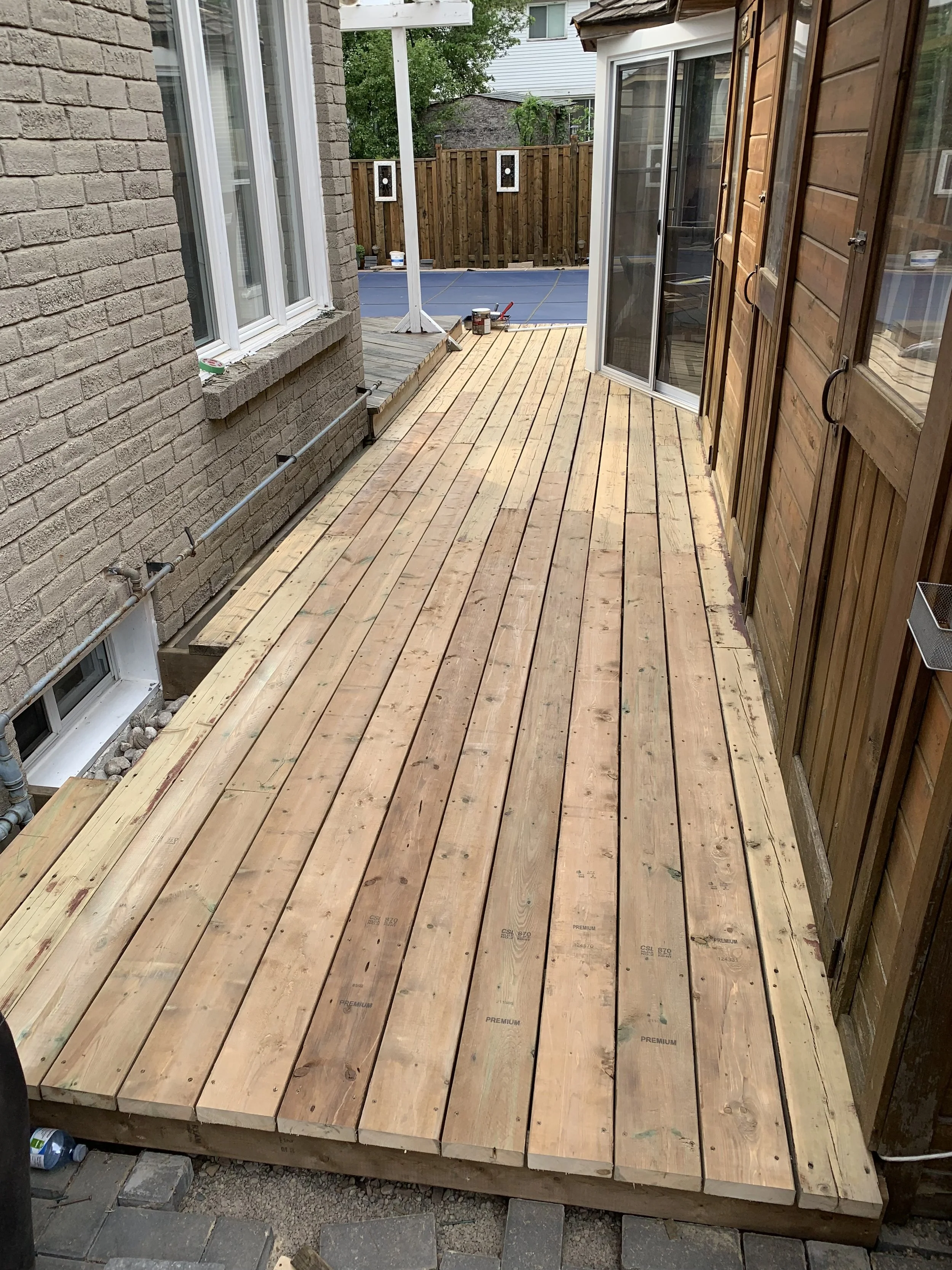 Newly constructed wooden deck with unfinished planks, adjacent to a brick house and a wooden shed, in a residential backyard.