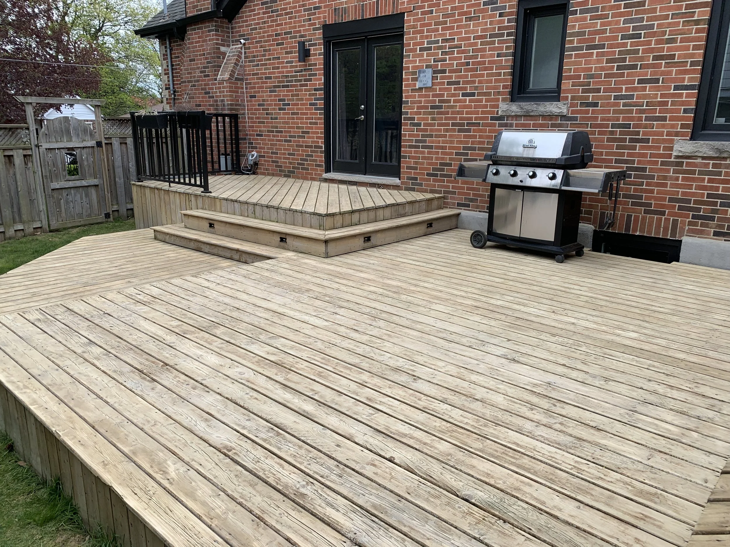 A wooden backyard deck with stairs leading up to a higher section, attached to a brick house with a sliding glass door. A black grill is on the right side of the deck, and a small black railing encloses part of the upper section. There is a grassy yard and a wooden fence in the background.