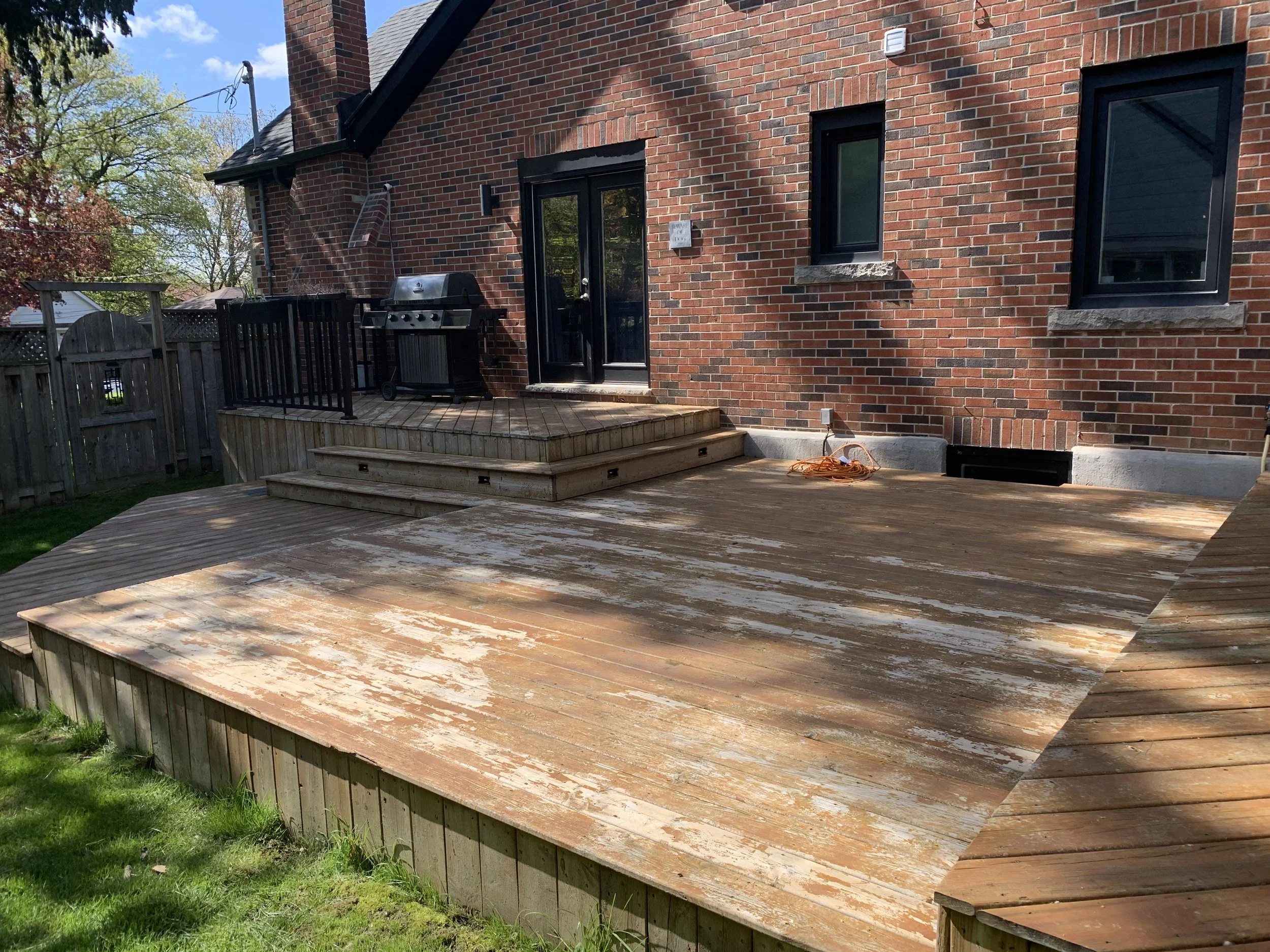 A backyard wooden deck attached to a brick house with two stained windows. The deck has a stairway with three steps and a black grill on the upper level. The lower part of the deck appears weathered and peeling.