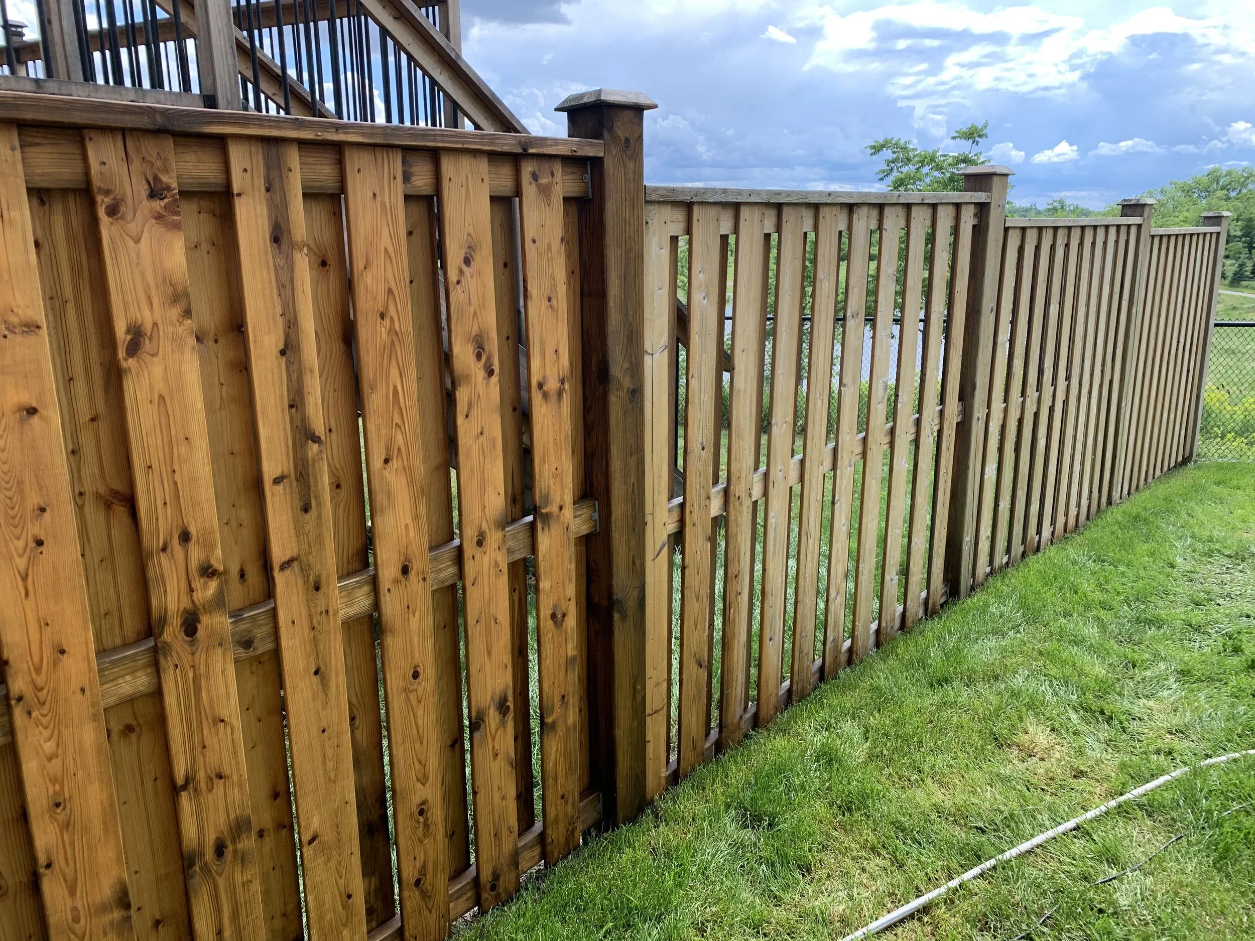 A long wooden fence with vertical planks and decorative caps, running along a grassy area, under a partly cloudy sky.
