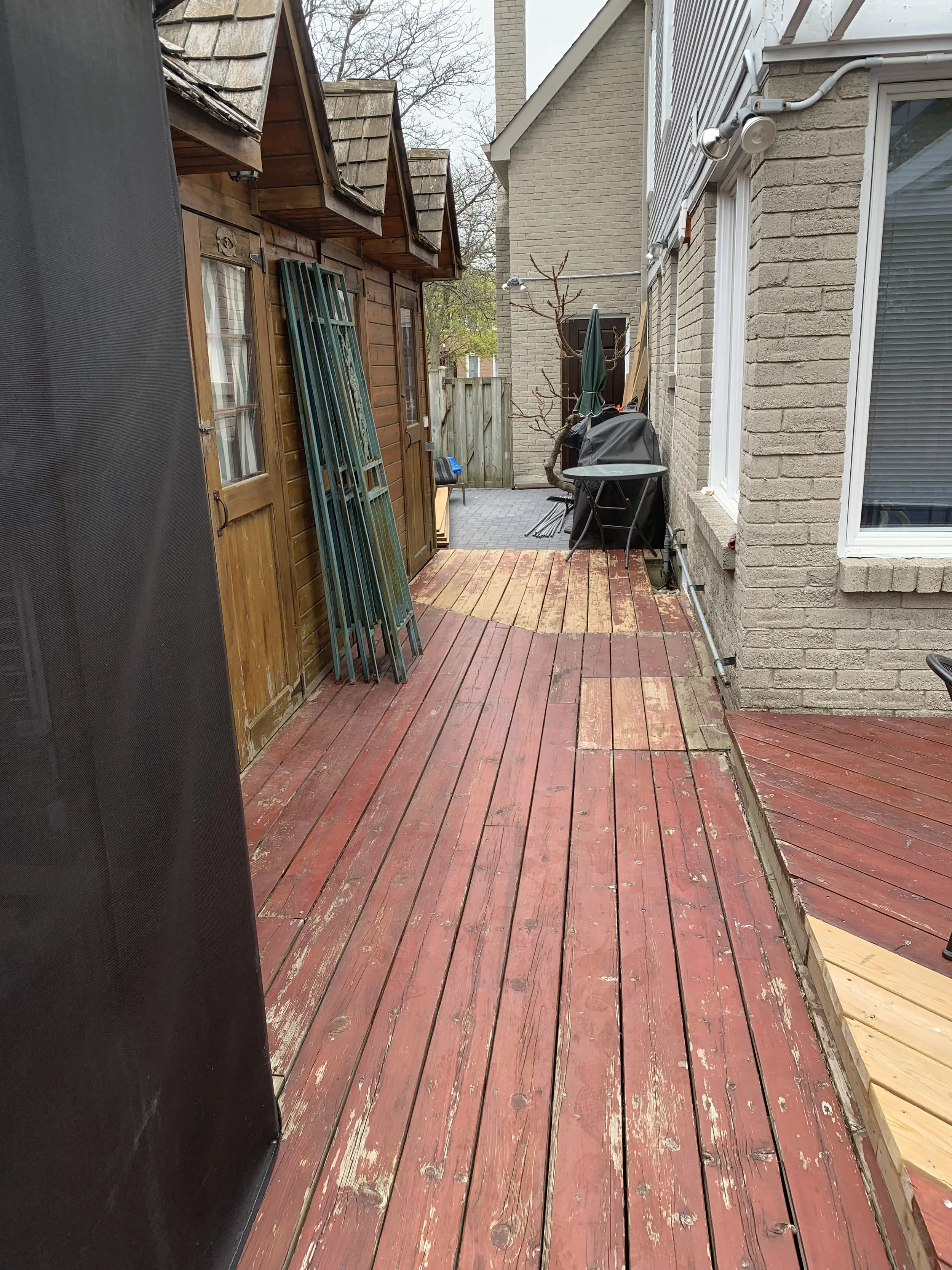 Backyard wooden deck with signs of weathering, a wooden shed with multiple roof sections on the left, and a brick house with a window on the right. An outdoor grill, furniture, and a small leafless tree are also visible.