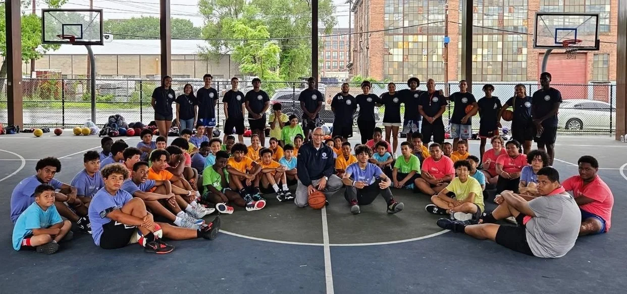 Group of children and coaches on an outdoor basketball court, some holding basketballs, with a chain-link fence and city buildings in the background.