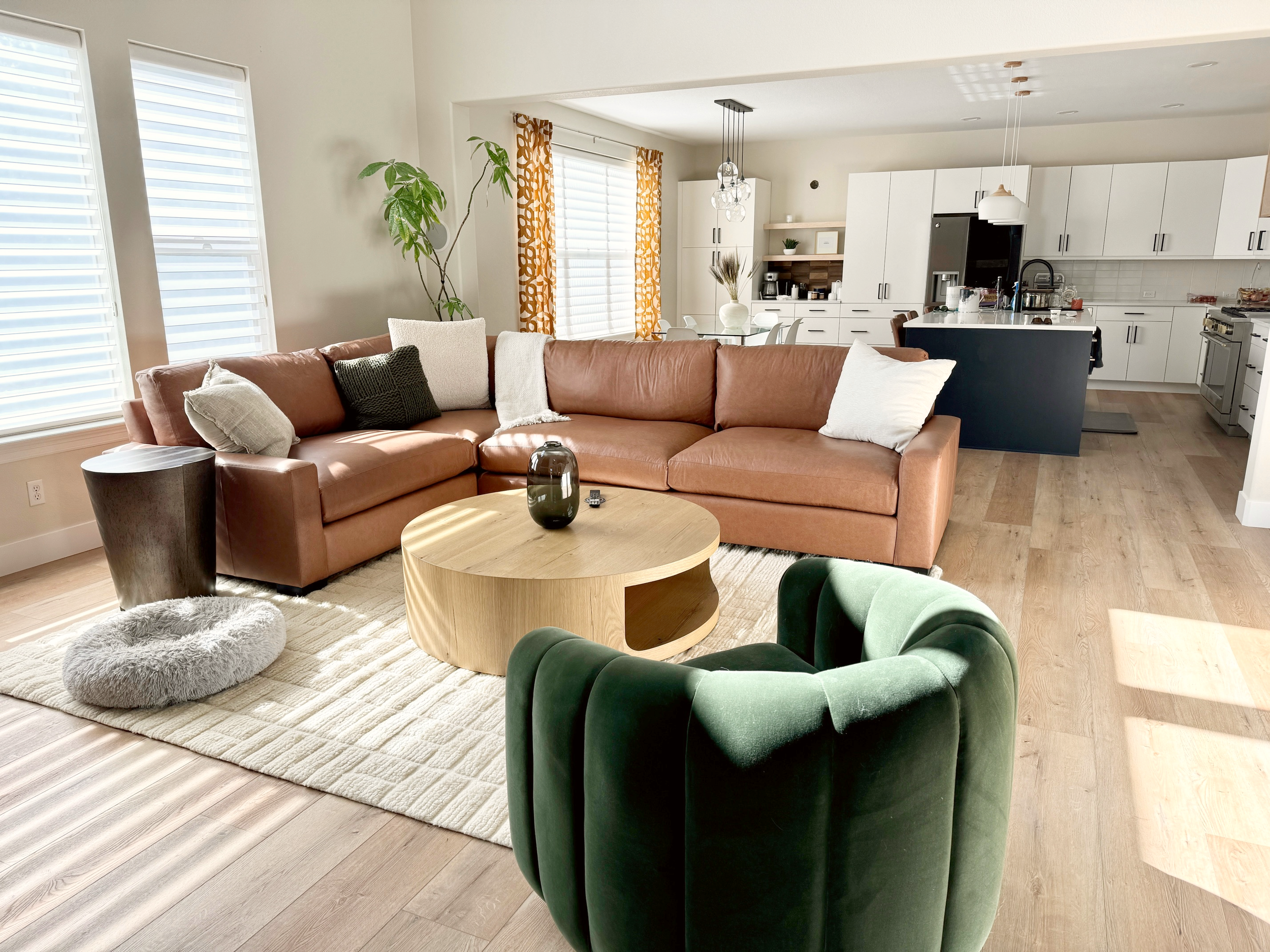 A cozy living room with a large brown leather sectional sofa, a green velvet armchair, a round wooden coffee table, and a cream area rug. The room is bright with sunlight streaming through window blinds, with a living room and kitchen visible in the background.