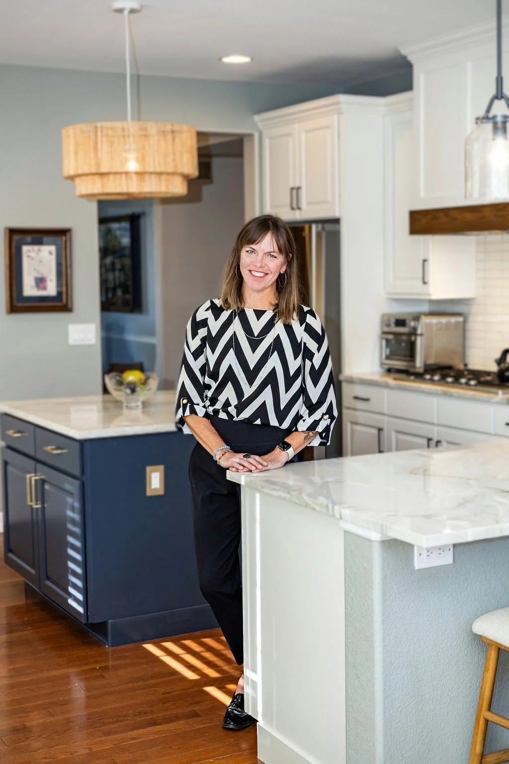 A woman standing in a modern kitchen, smiling, wearing a black and white zigzag patterned blouse and black pants.