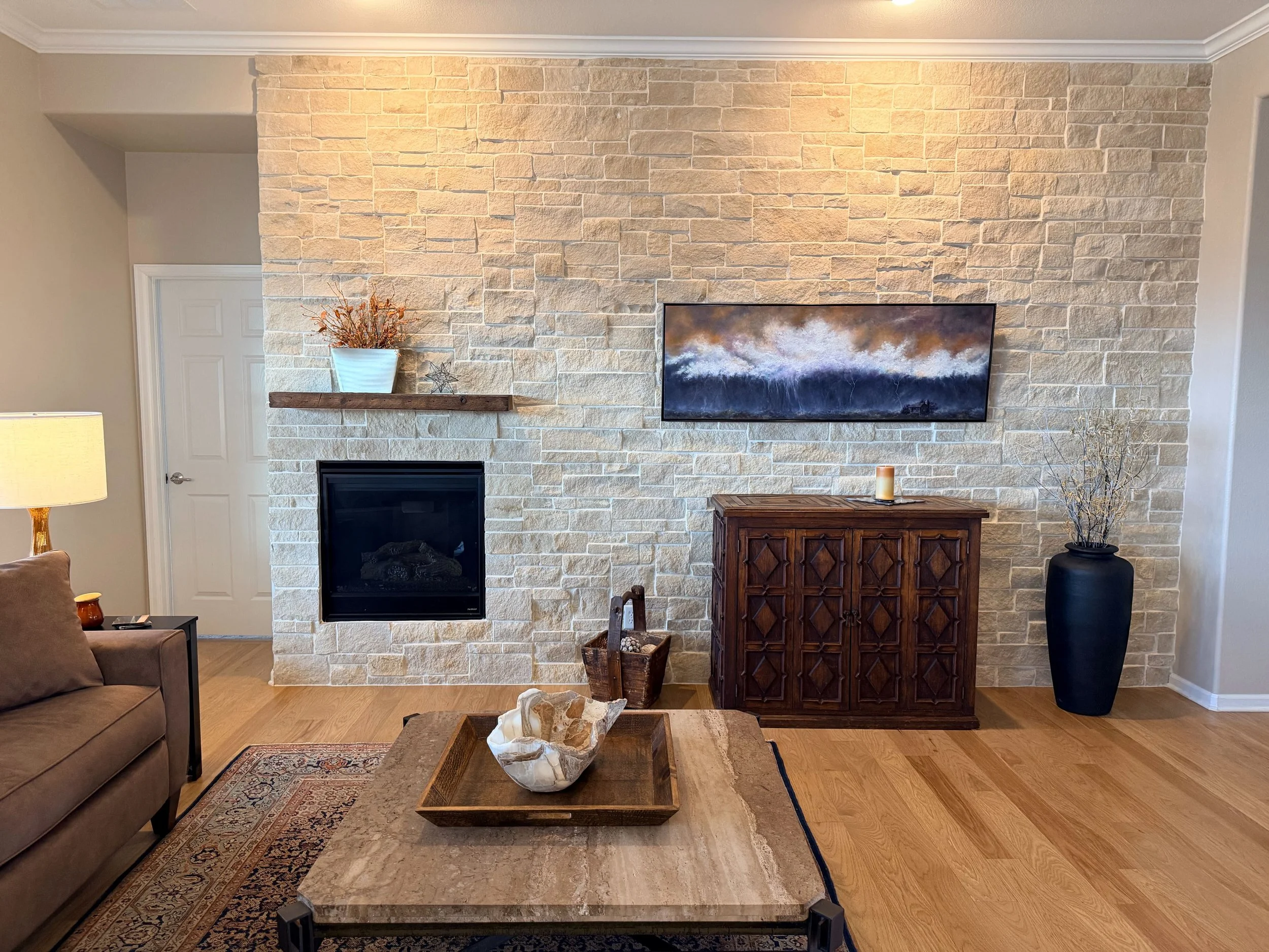 Living room with beige brick fireplace, wooden mantel, wall-mounted TV, and decorative items including a large black vase with branches, a wooden cabinet, a stone bowl on a coffee table, and a area rug.