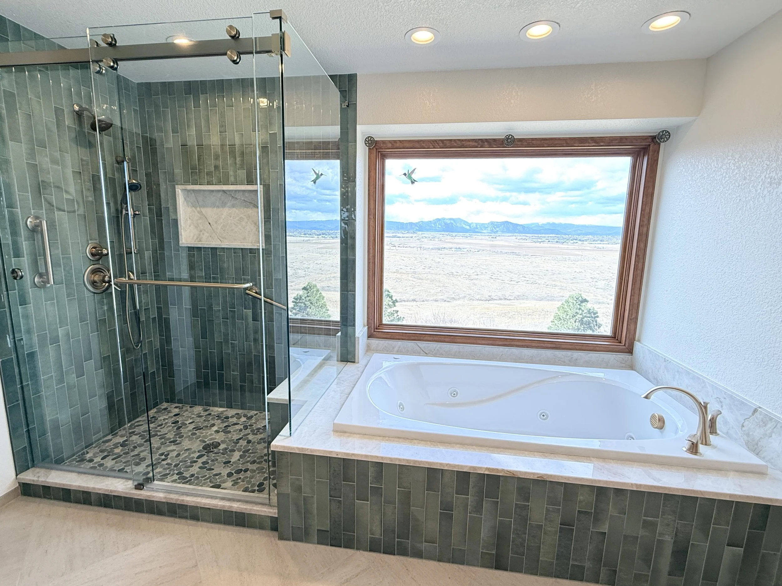Bathroom with large window overlooking a mountain landscape, featuring a glass-enclosed shower with dark green tiles and a pebble floor, and a white bathtub with marble surround.