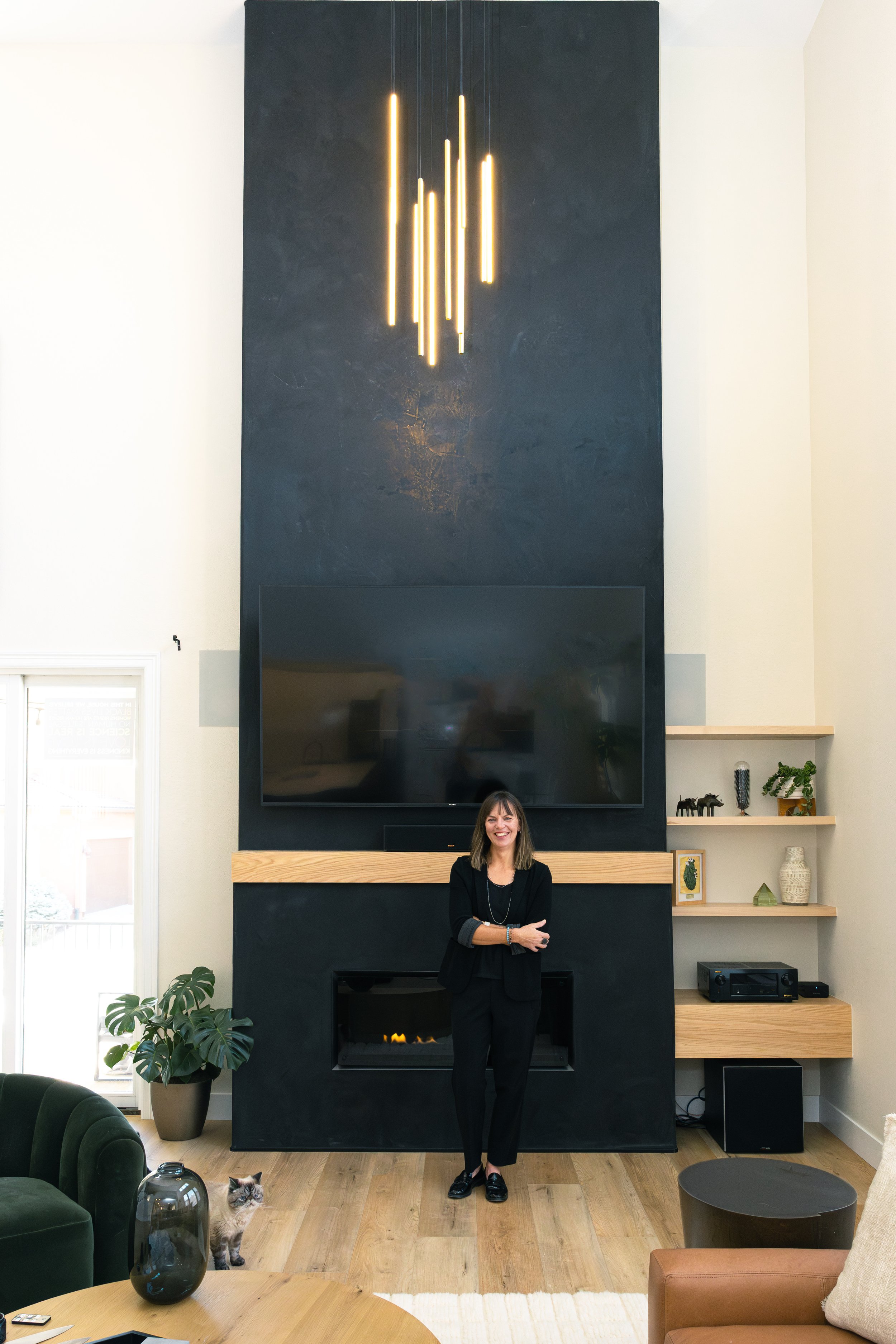A woman standing and smiling in a modern living room with a black feature wall, a large TV, a fireplace, and wooden shelves with decor items, a cat, and a large potted plant.