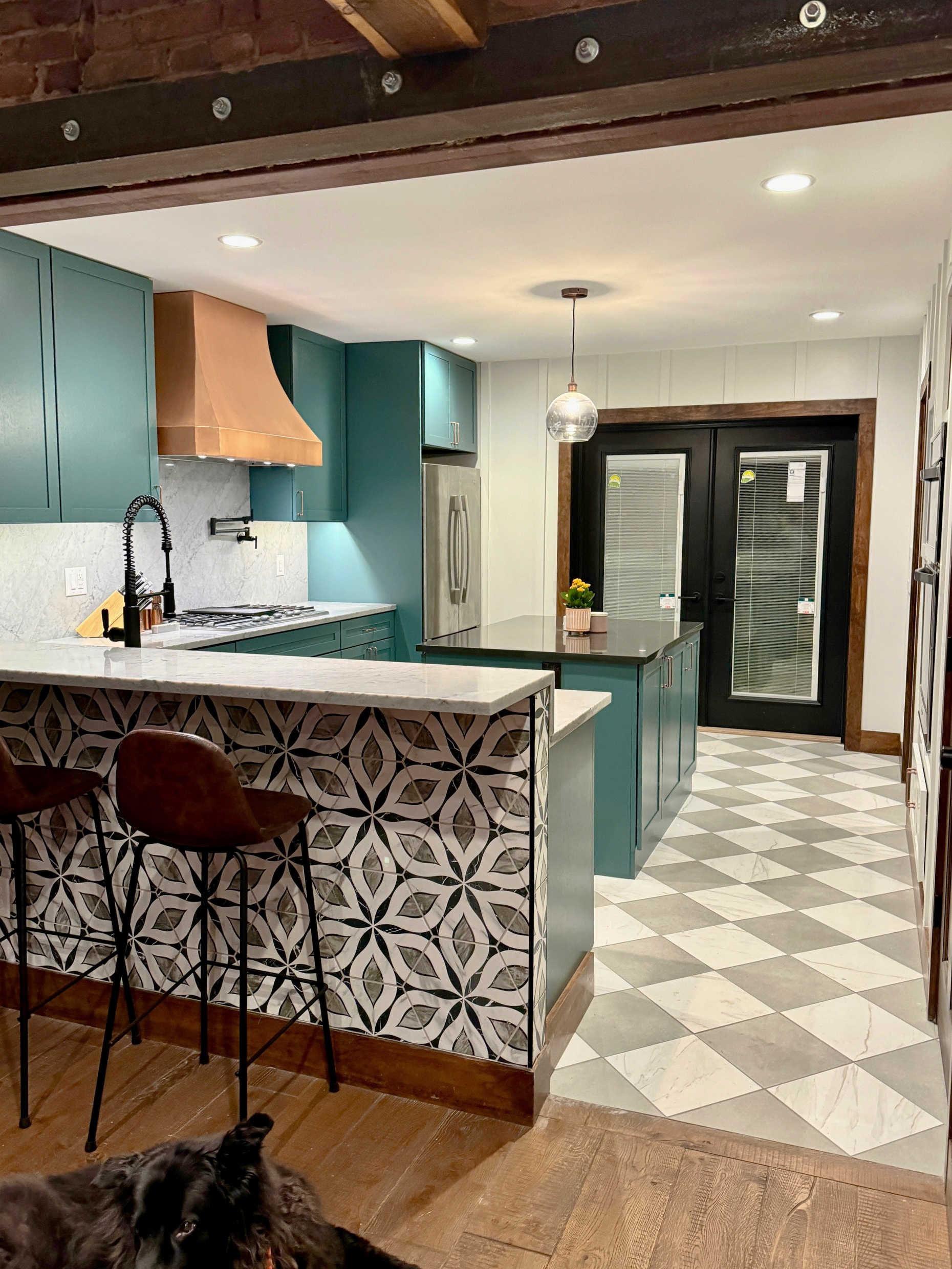 Modern kitchen with teal cabinets, copper range hood, patterned tile on the front of the island, and a black and white diamond-patterned floor. There is a hanging light fixture above the island, a small potted plant, and double glass doors leading outside. Part of a black dog is visible in the foreground.