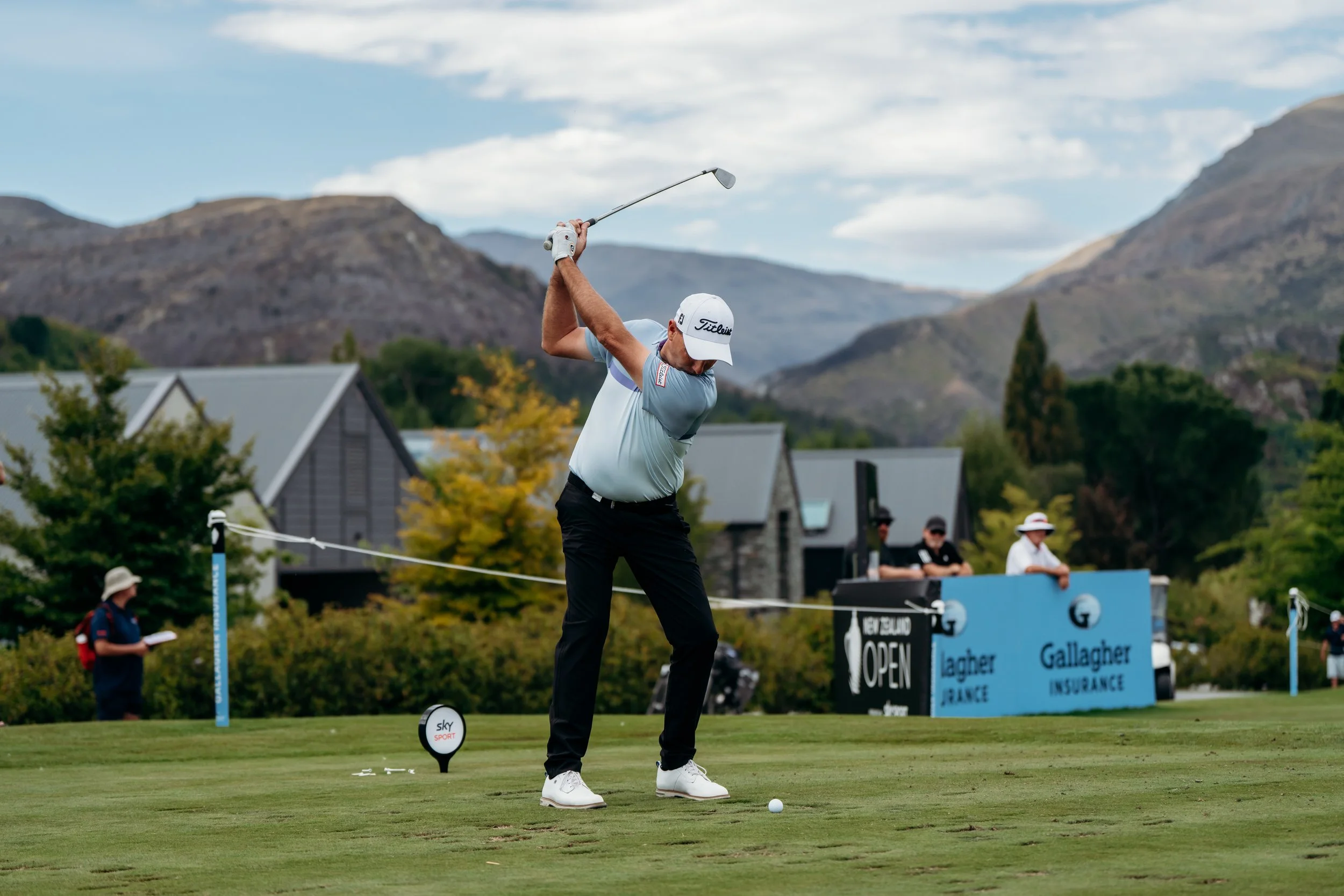A golfer in a pale blue shirt and black pants is taking a shot on a golf course with mountainous terrain in the background. He is swinging a golf club and watching the golf ball on the ground. Several people are observing in the background, some holding clipboards. The setting appears to be during a professional golf event.