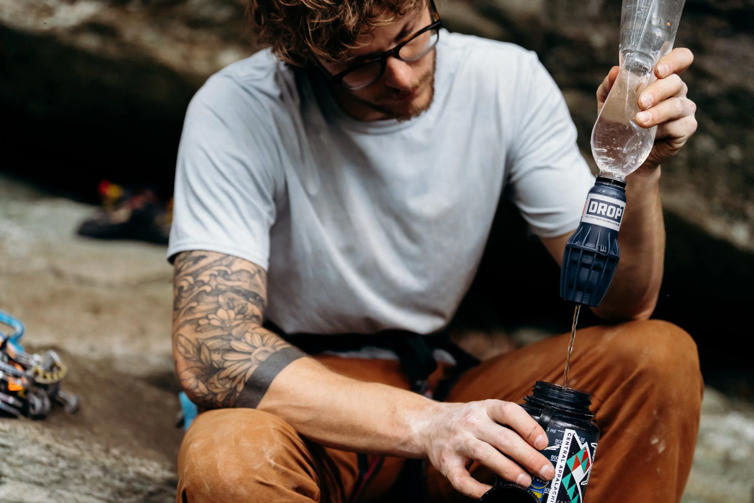 Man with glasses and tattoos sits on the ground outdoors, pouring liquid from a plastic water bottle into a black container.