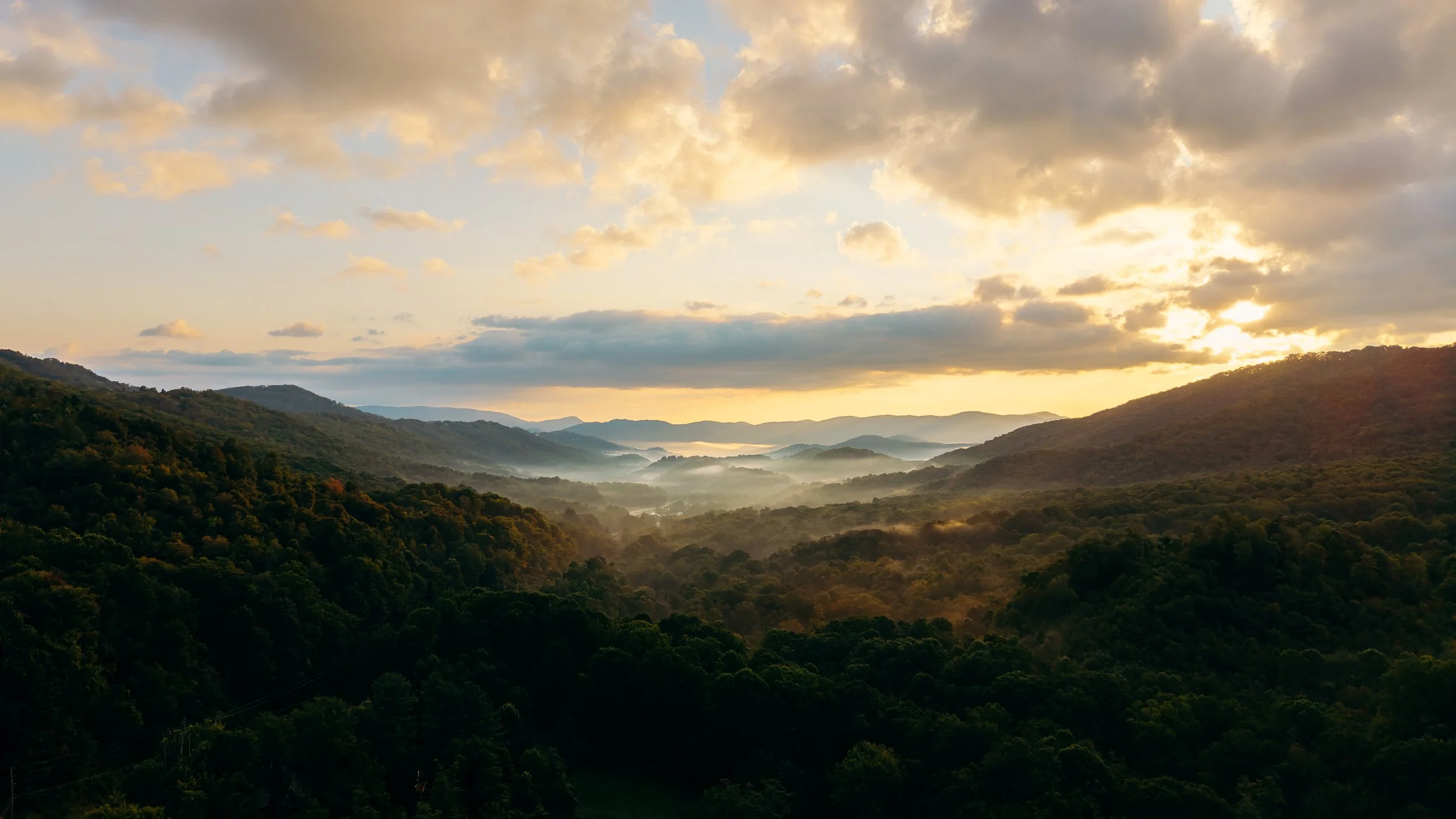 Sunset over a mountain valley with misty hills and green forested slopes.