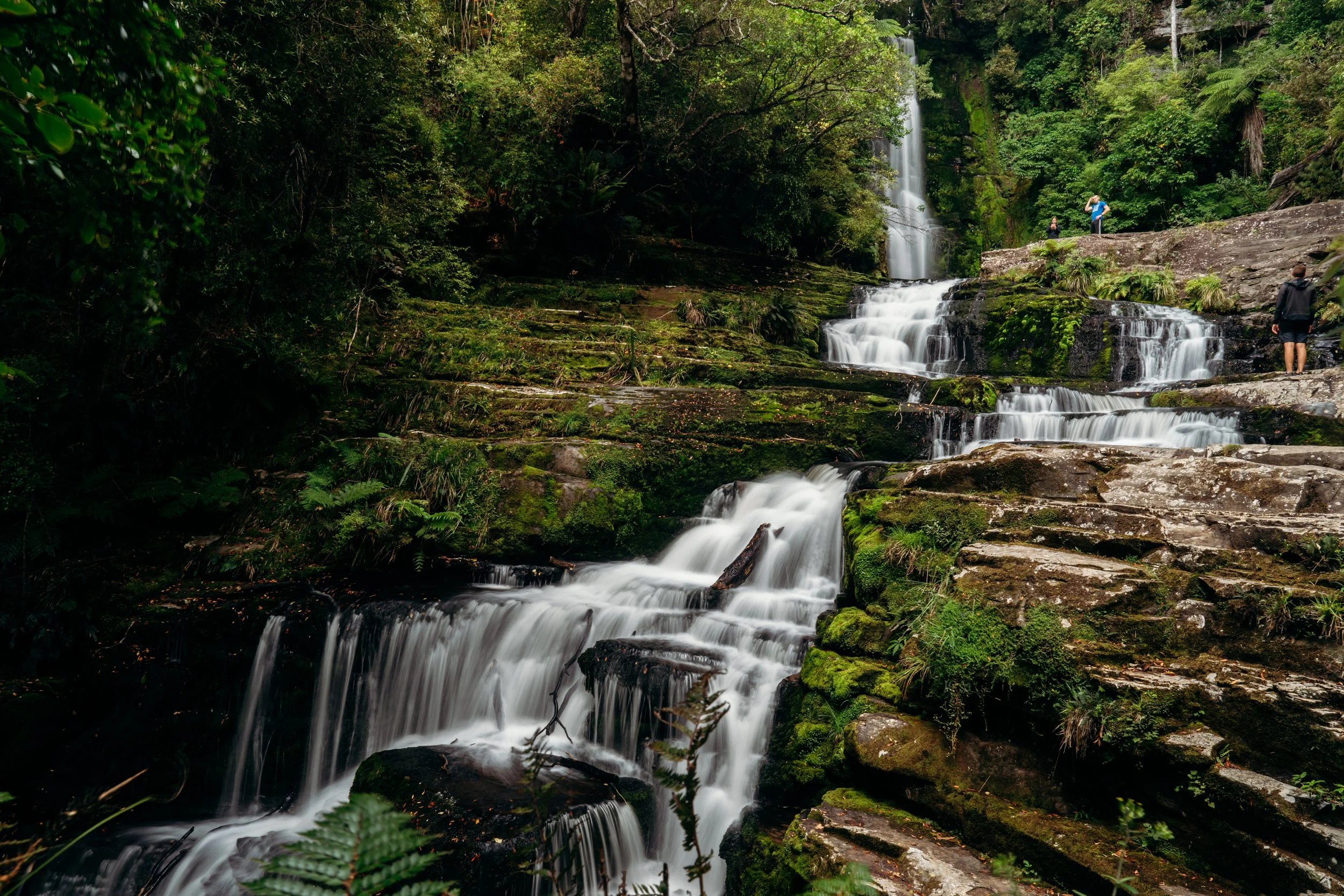 A multi-tiered waterfall cascading down moss-covered rocks in a lush forest with three people exploring near the waterfall.