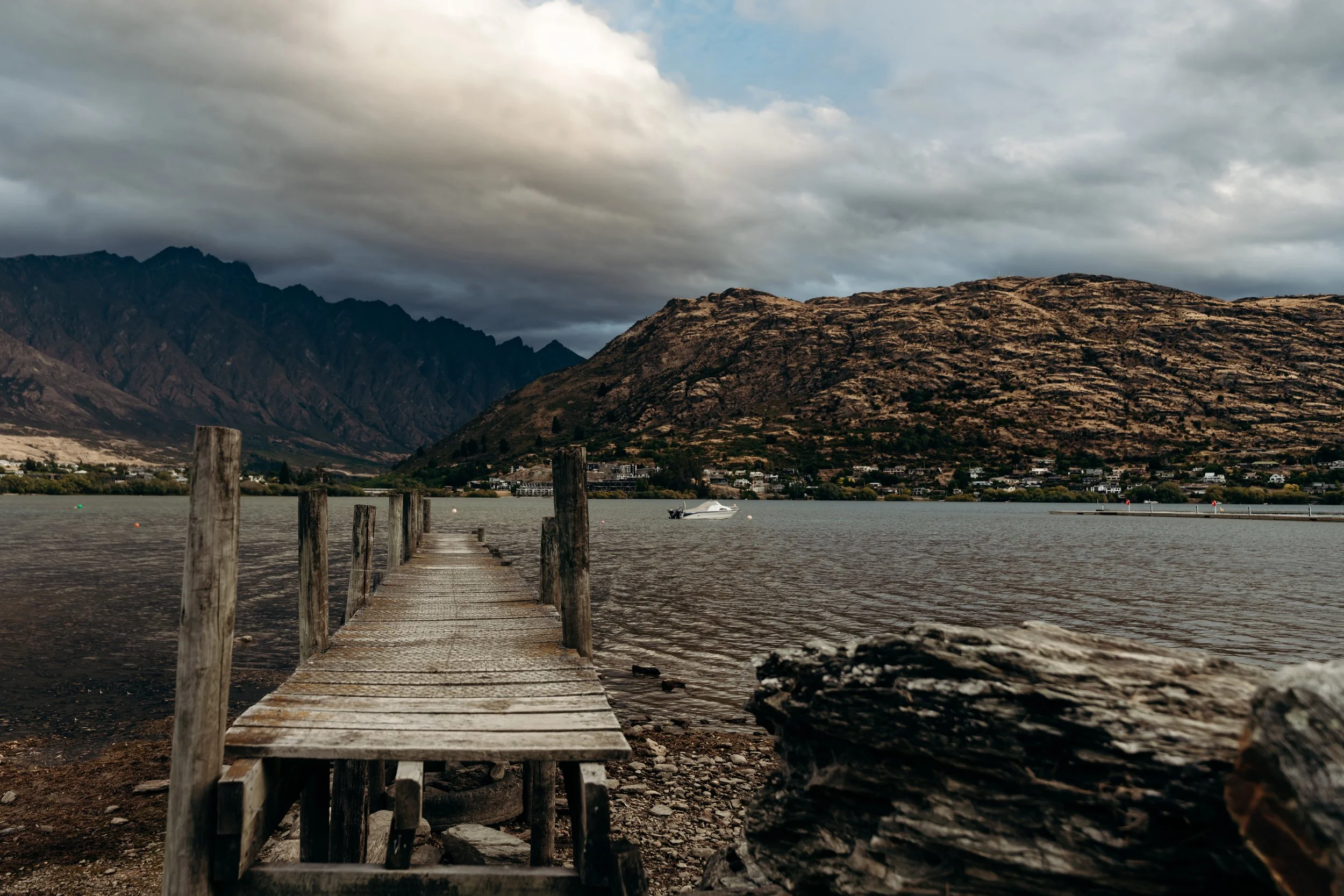A wooden dock extends into a body of water with mountains in the background and cloudy skies overhead.