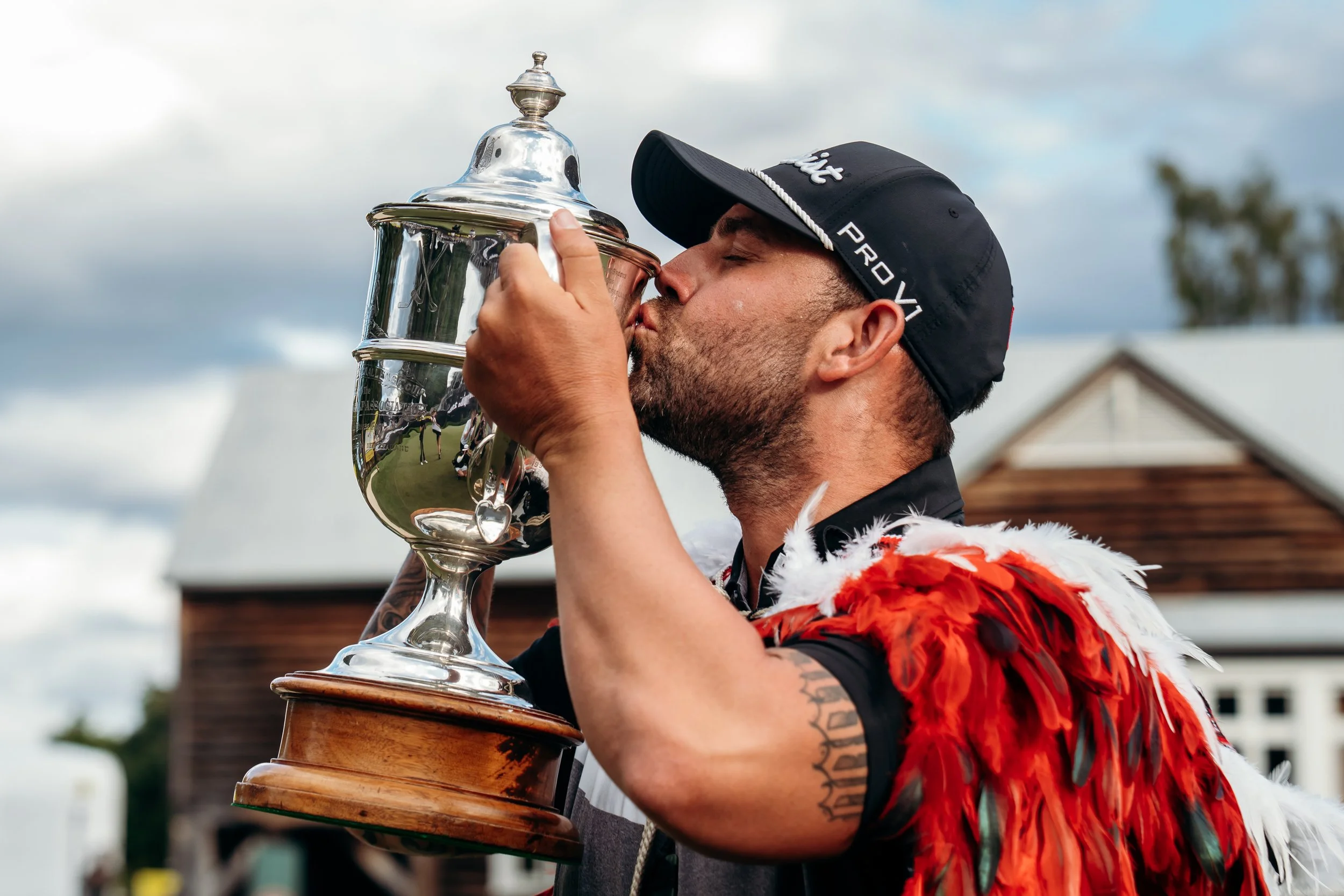 A man wearing a black cap and a colorful feathered vest is kissing a shiny silver trophy. The man has tattoos on his arm, and the background shows a cloudy sky and buildings.