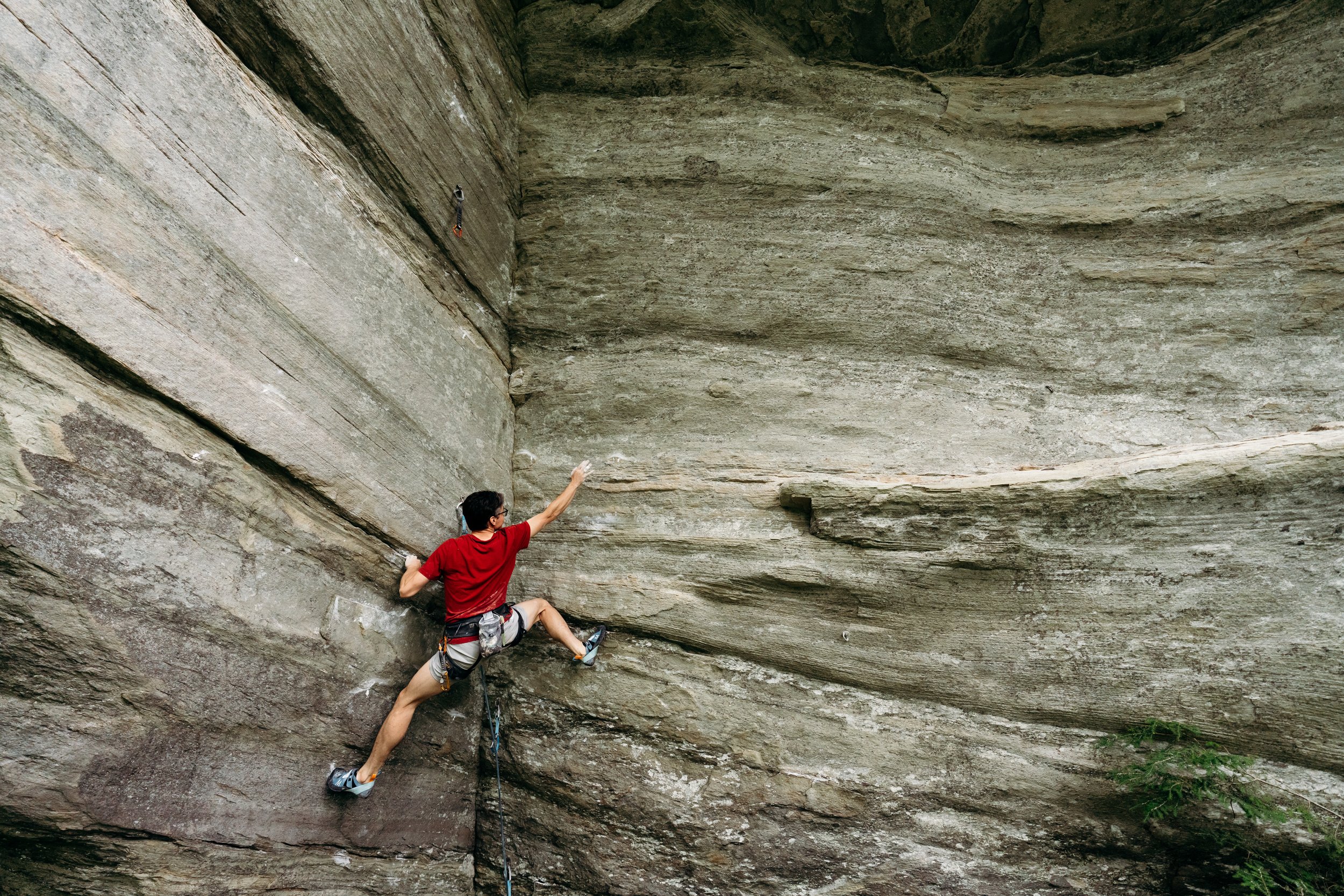 A man rock climbing on a steep outdoor rock face, dressed in a red shirt and climbing gear, reaching with his right hand while holding onto the rock with his left, with trees at the base.