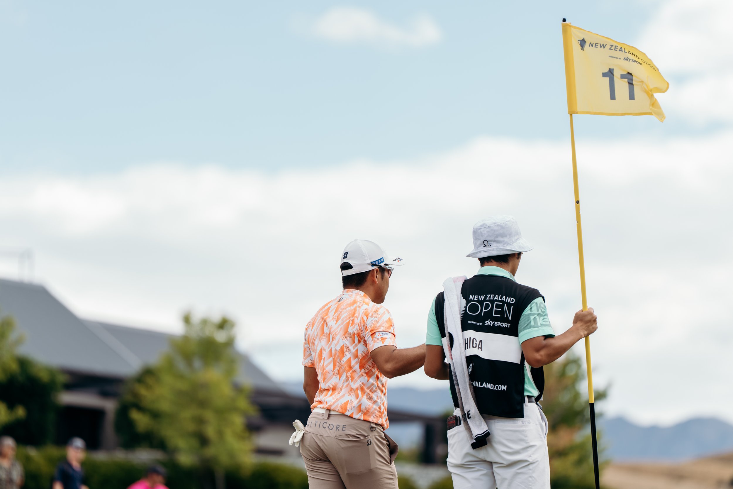 Golfers on a course, one holding a flag with the number 11 and 'New Zealand Open' written on it, with a partly cloudy sky overhead.