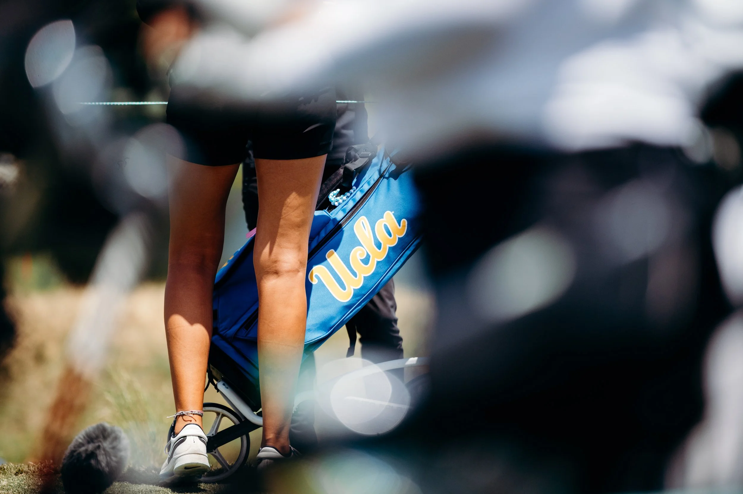 A person wearing athletic shorts and sneakers riding a bicycle on a grassy area, carrying a blue bag with the word "Nela" on it.