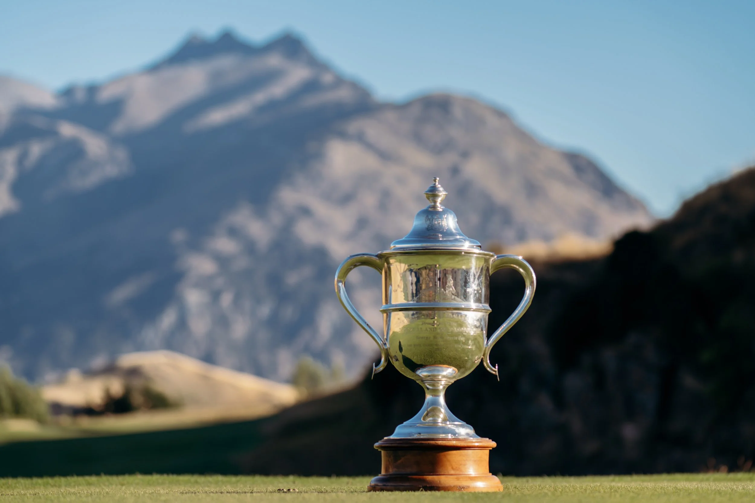 A large silver trophy with a lid, handles, and a reflective surface, placed on a wooden base on a golf course, with mountains in the background under a clear blue sky.