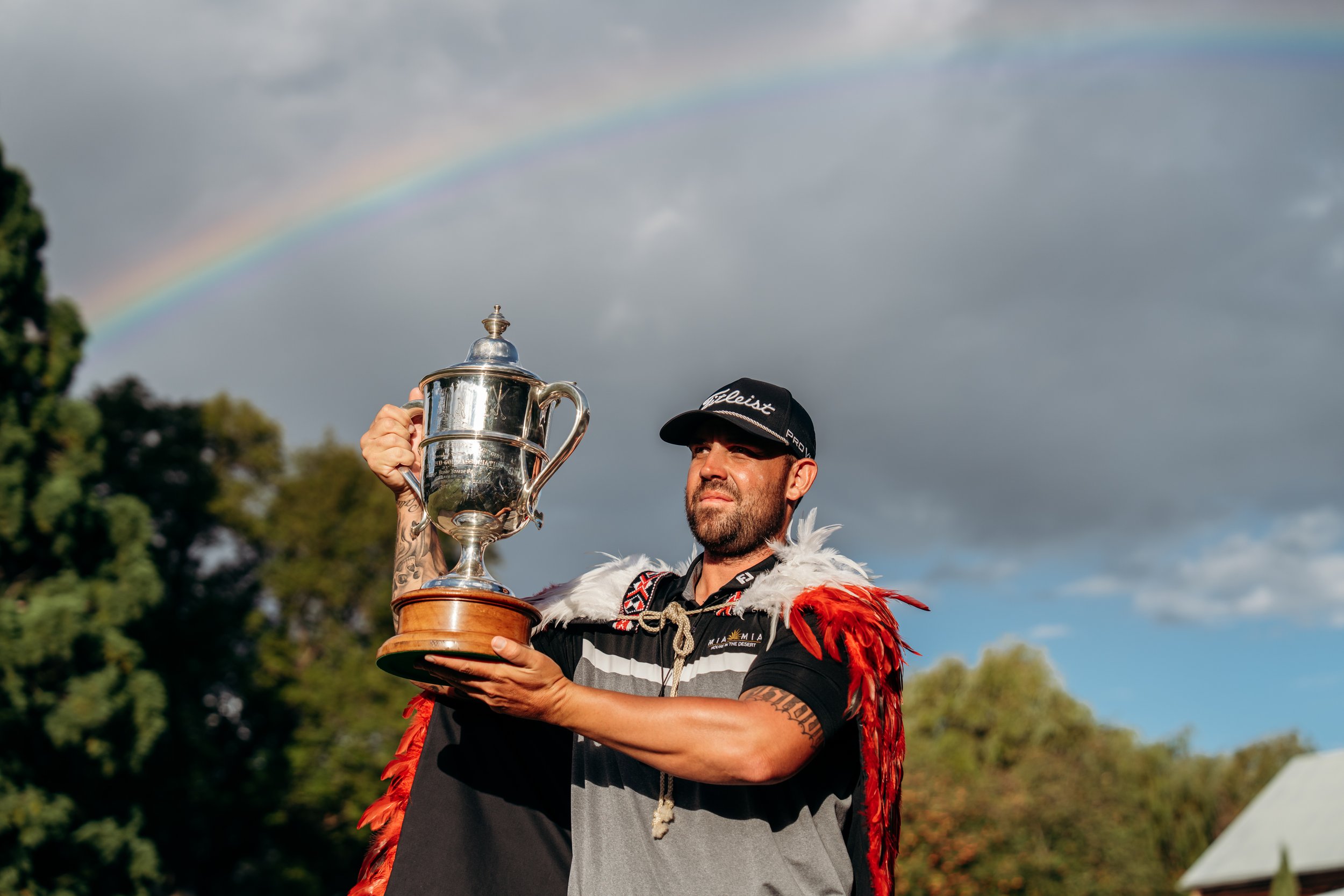 A man with a beard, wearing a black cap and sportswear, holding up a silver trophy in his right hand, with a rainbow visible in the cloudy sky behind him.