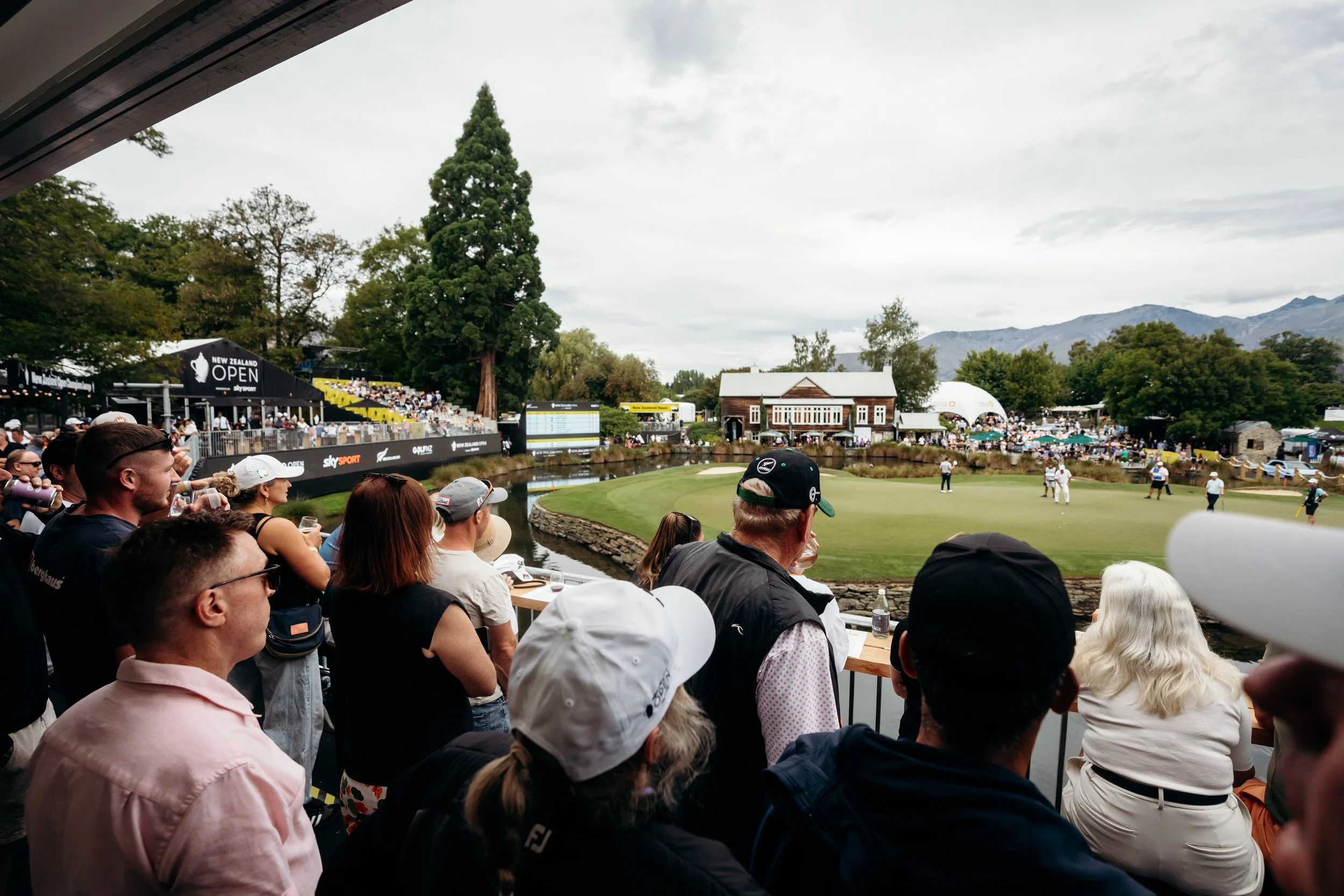 View of a golf course filled with spectators, banners, and a backdrop of trees and mountains during a tournament.
