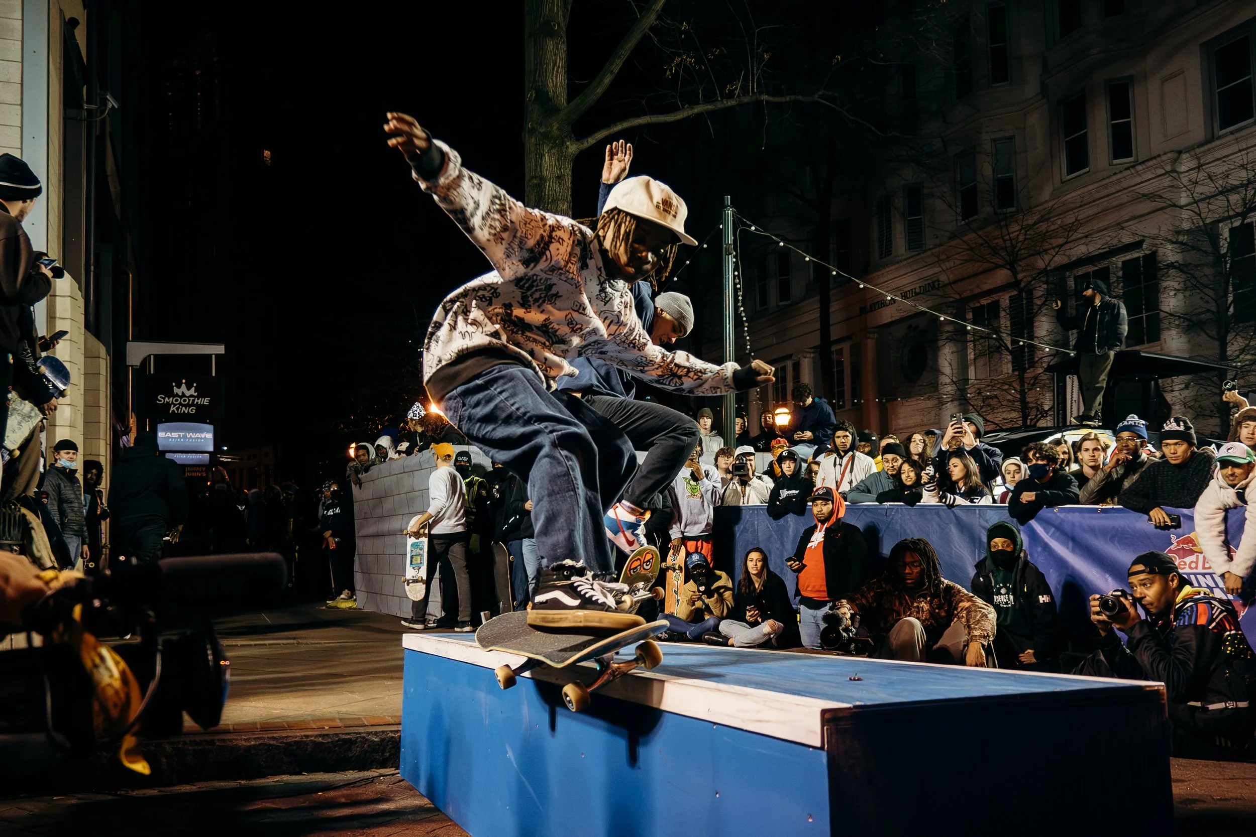 A skateboarder performing a trick on a ledge at an outdoor event at night, with a crowd watching and taking photos. The scene includes streetlights, trees, and buildings in the background.