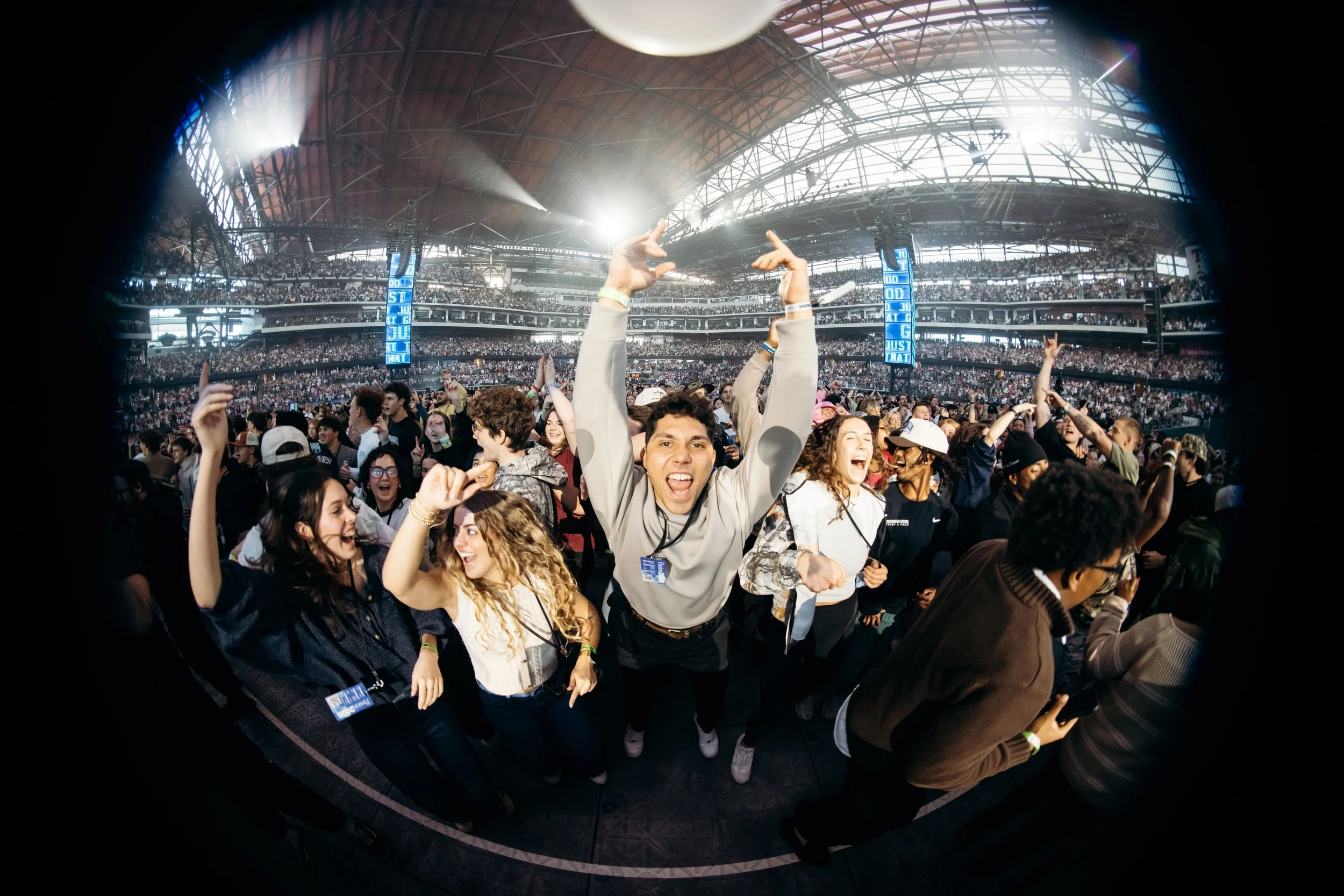 A large crowd of people is attending an indoor event, possibly a concert or sports game, in a stadium with a high, curved ceiling. Many are cheering, smiling, and raising their hands, with some making peace signs or pointing upward.