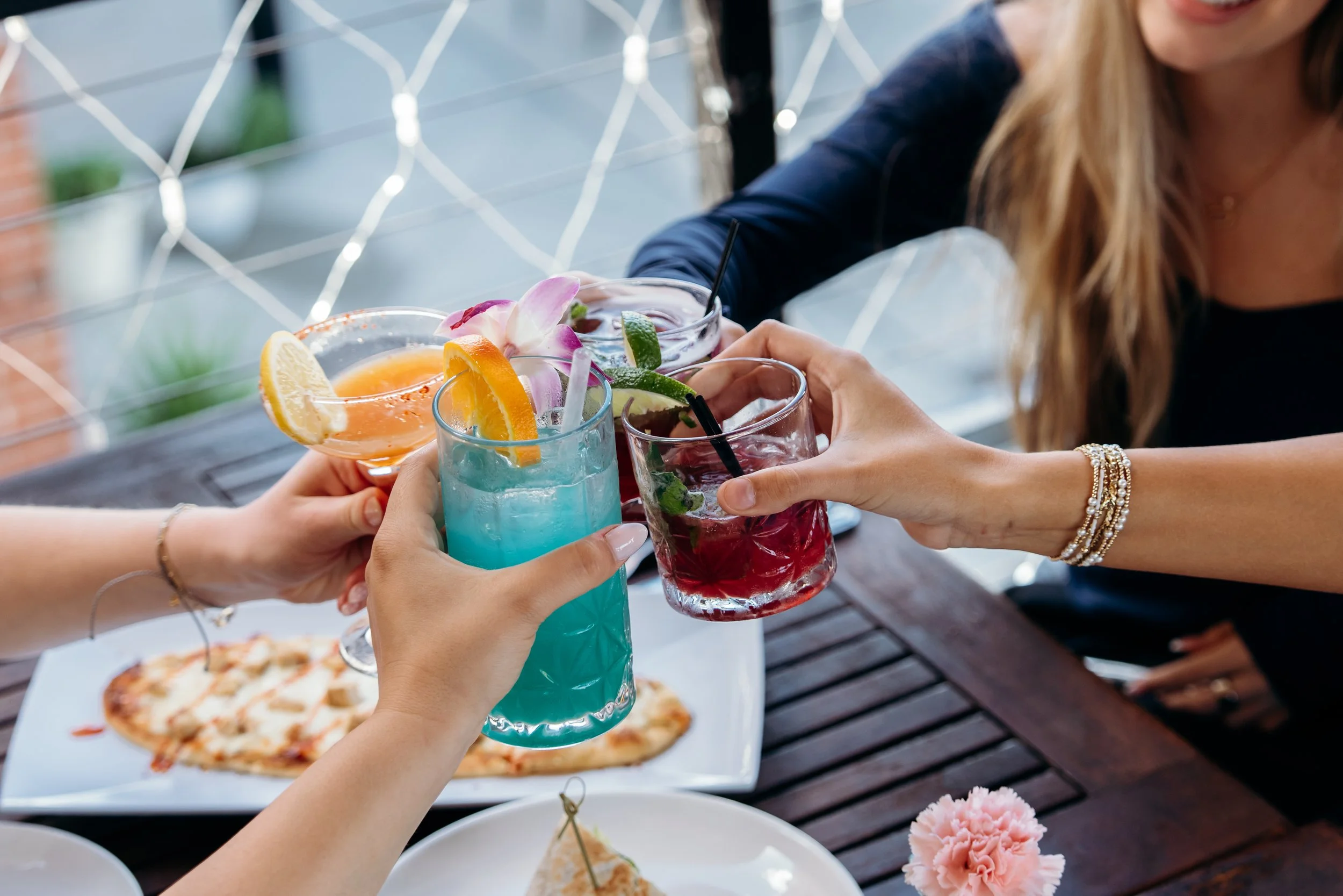 A group of people raising colorful cocktails in a toast at an outdoor dining table, with pizza and pink flowers visible on the table.