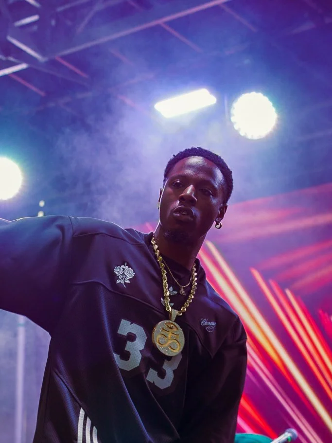 A young man with jewelry and earrings in a dark sports jersey on stage with colorful lights and laser effects in the background.