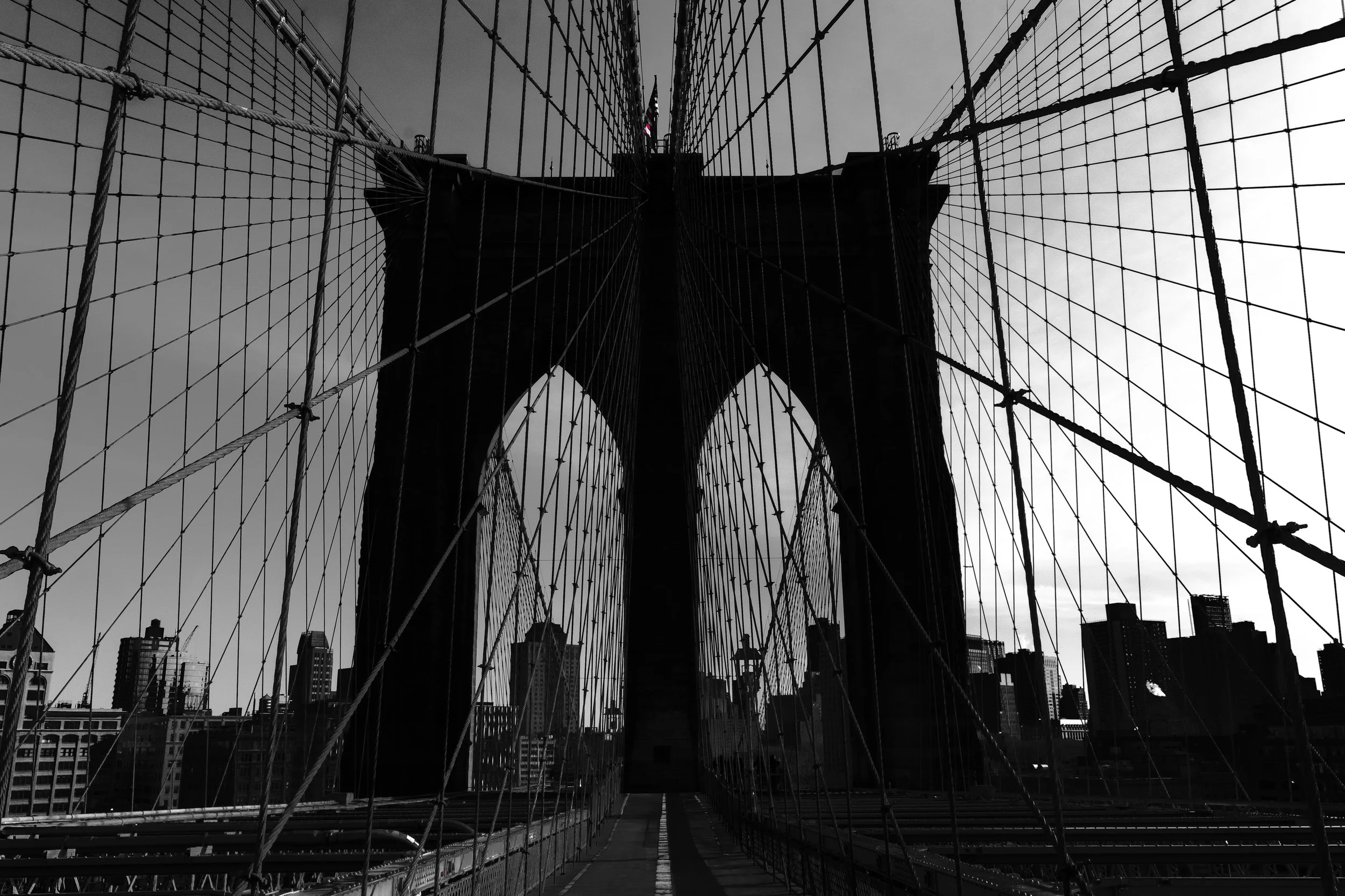 A black and white photo of the Brooklyn Bridge in New York City, taken from the pedestrian walkway looking up at the bridge's towers and suspension cables with city buildings in the background.