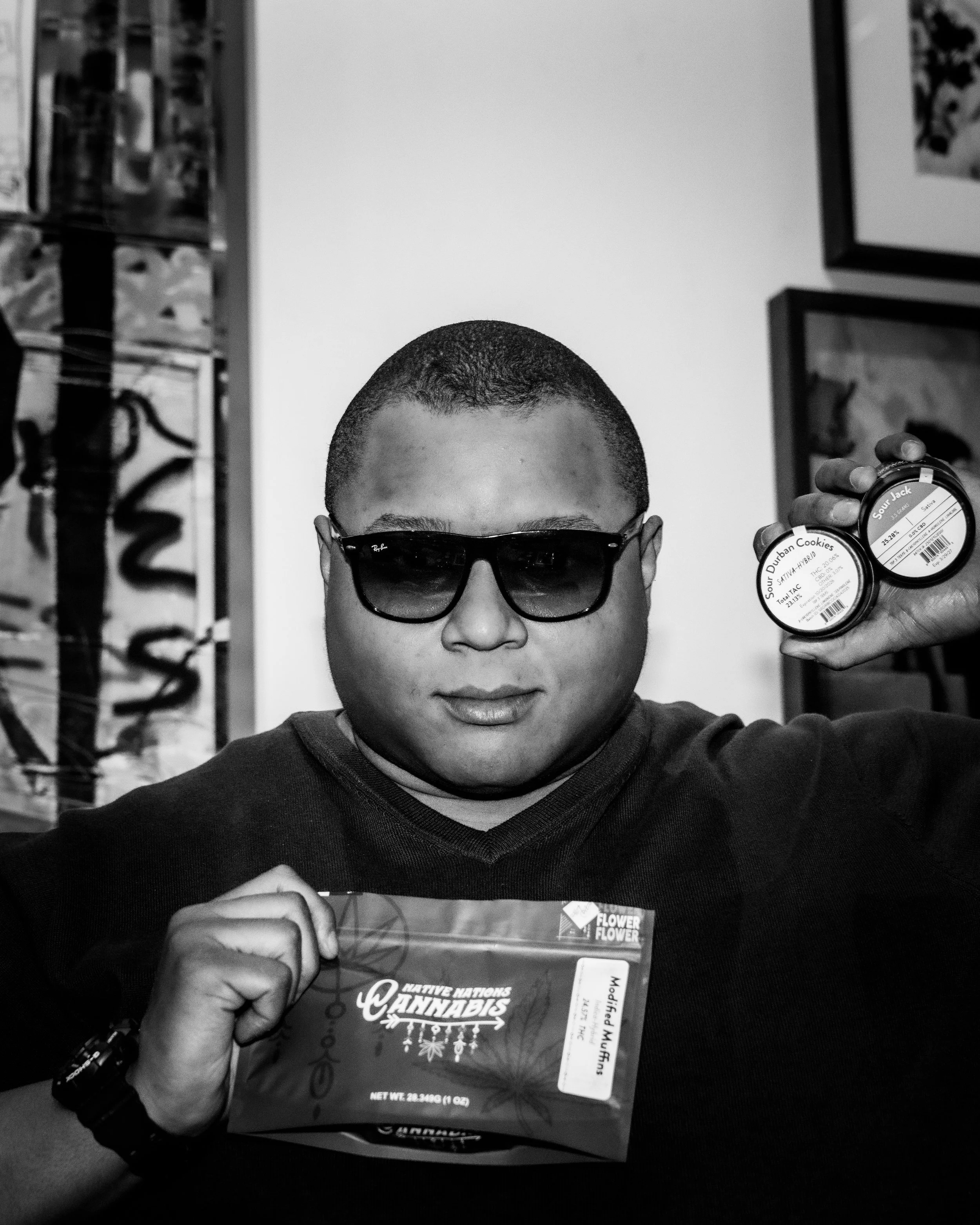 A man wearing sunglasses is holding a cannabis-themed chocolate bar in front of him, and in his other hand, he shows two containers of cannabis edibles. The background includes a room with framed pictures and a display case.