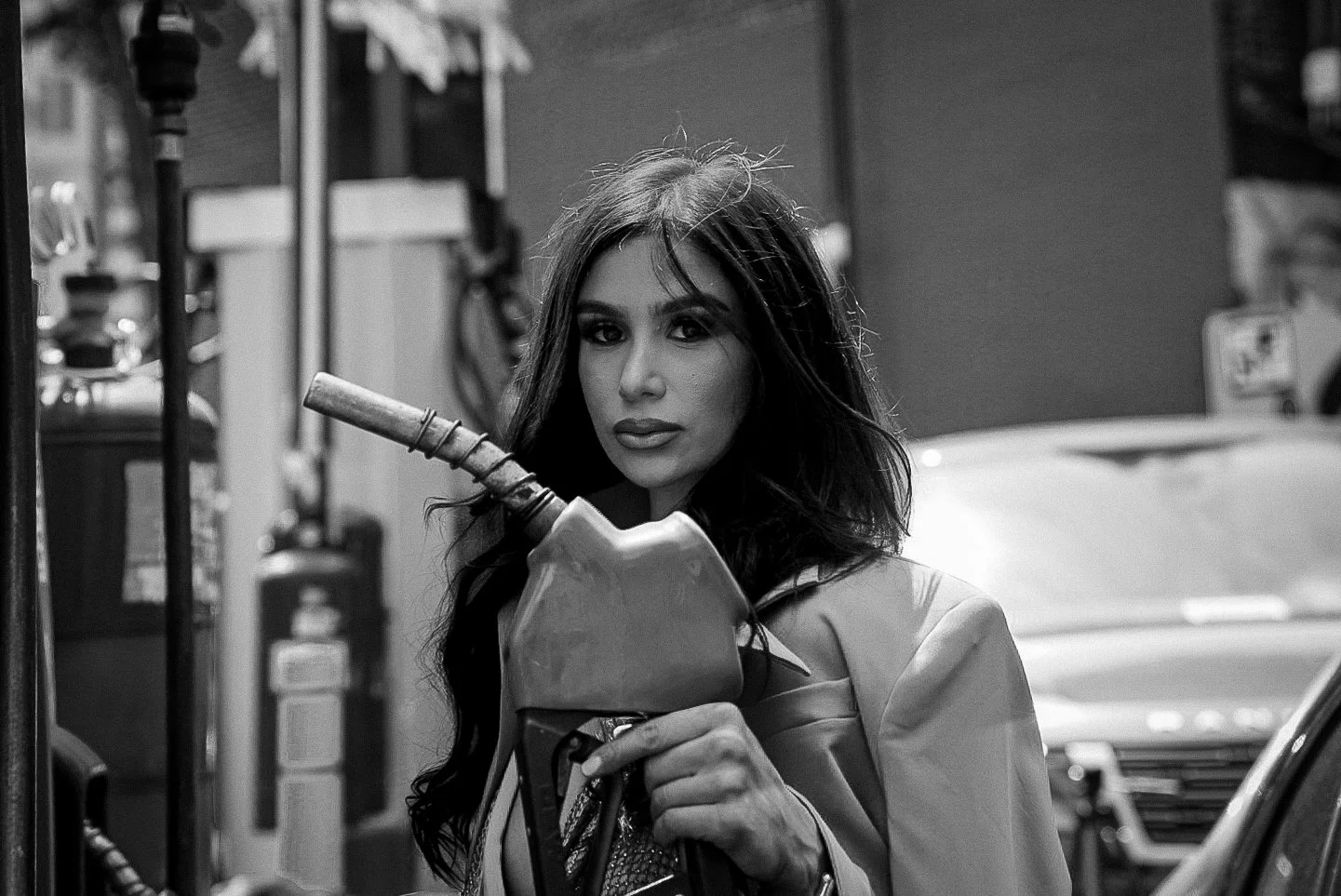Black and white photo of a woman with long dark hair holding a gasoline pump nozzle in a gas station.