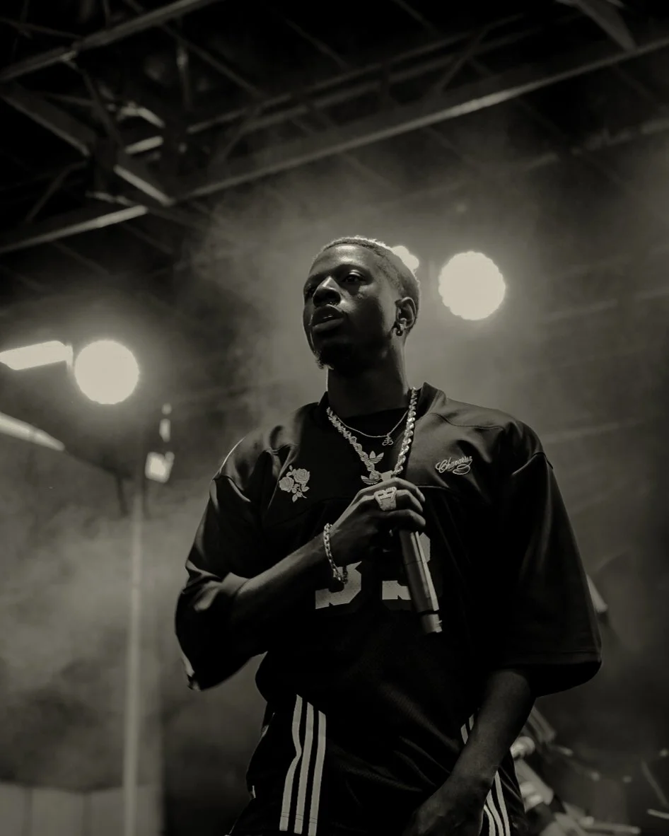 A Black man holding a microphone on stage, dressed in a dark sports jersey and jewelry, under bright stage lights, with a smoky background and ceiling structure visible.