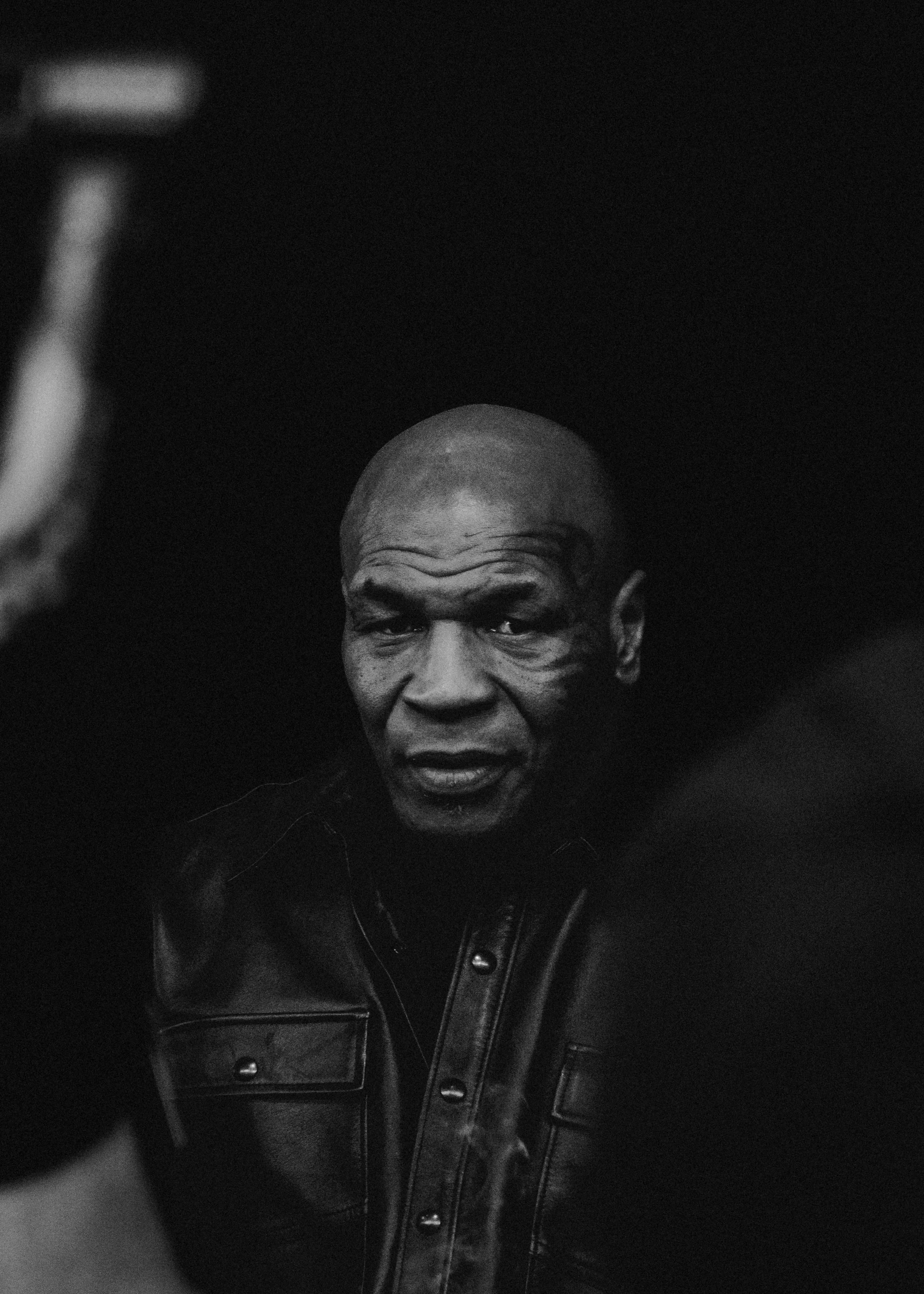 Black and white photograph of an older man with a bald head, wearing a leather jacket, looking directly at the camera with a serious expression.