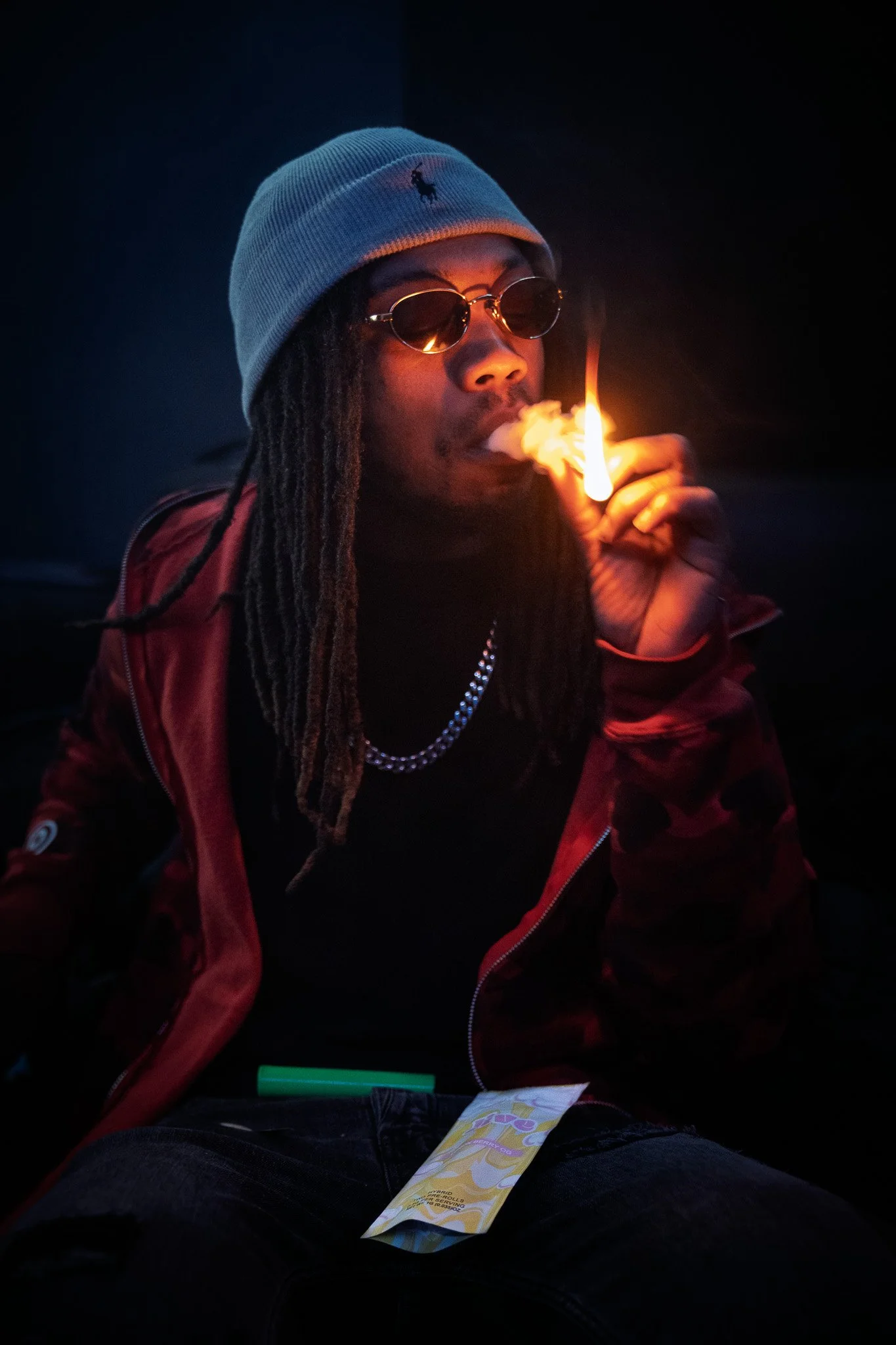 A man with dreadlocks wearing a gray beanie, sunglasses, a red jacket, and a black shirt, smoking while sitting in a dark setting with a lit joint in his hand.